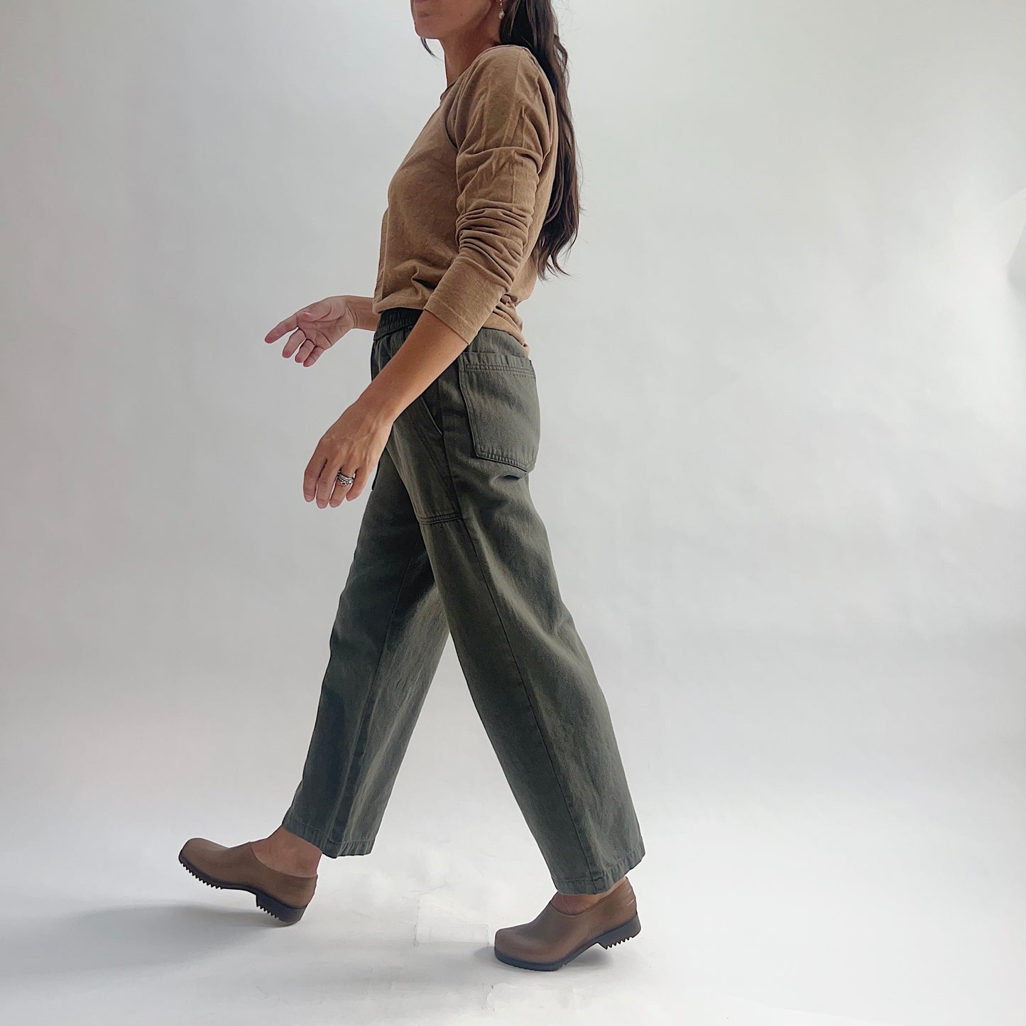 Side profile of model walking in olive green wide-leg pants with a back patch pocket visible, styled with a tan long-sleeve top and brown clogs.
