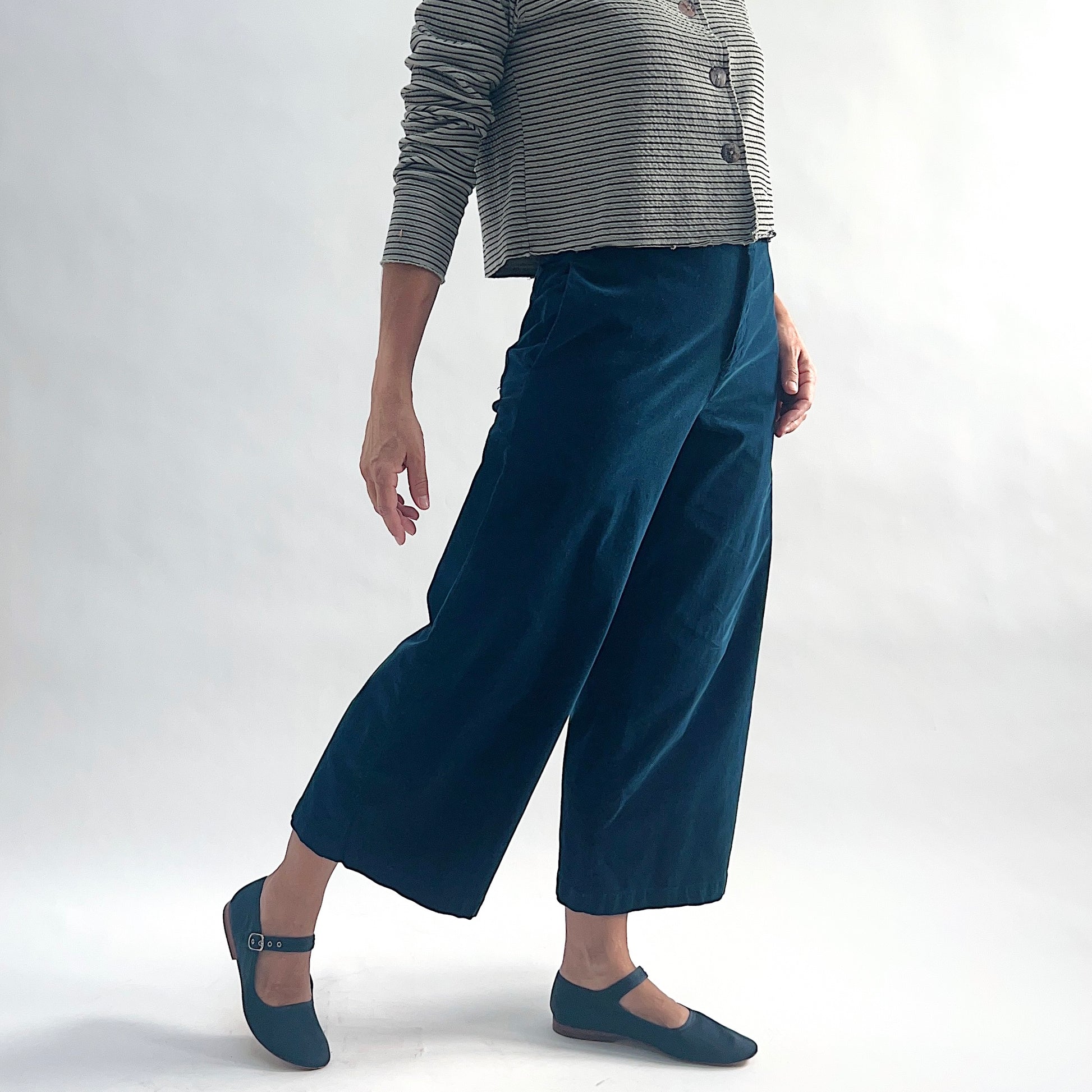 Three-quarter front view of model wearing deep teal mini cord straight crop trousers, leg extended mid-stride, showing the wide straight leg, flat front, and cropped length, styled with the matching white and black stripe asymmetrical cardigan and teal mary jane flats against a white background.

