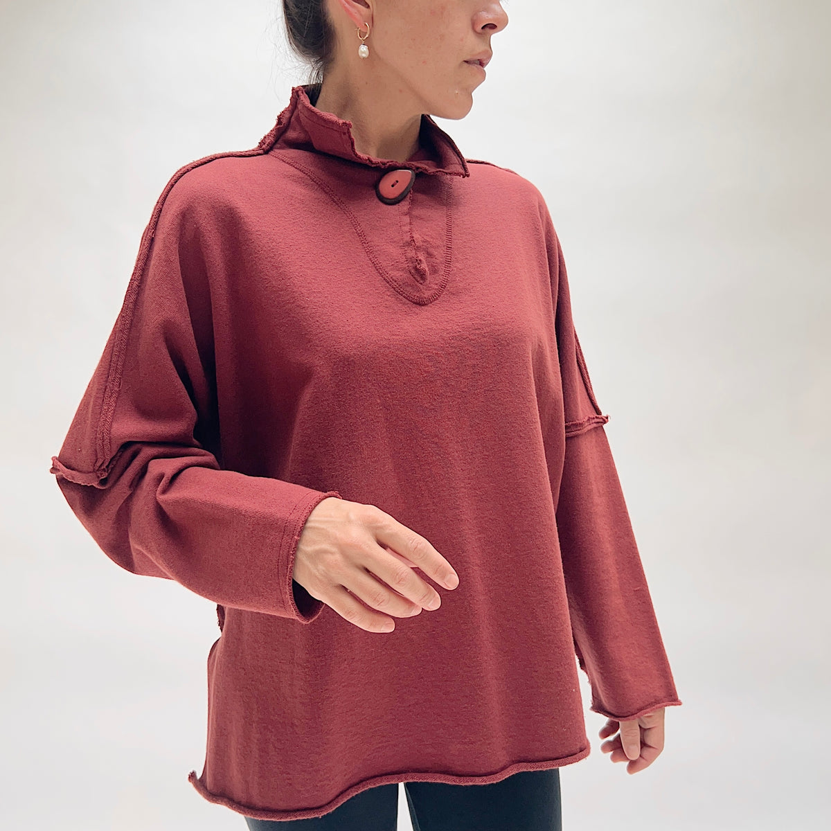 Model wearing the Eleven Stitch Long Rib Out Funnel Neck Slub Sweatshirt in Berry from a three-quarter angle, showing the ruffled funnel neckline with single large button, exposed raw rib seams at the shoulder and sleeve seams, and oversized drop-shoulder silhouette in deep berry slub cotton
