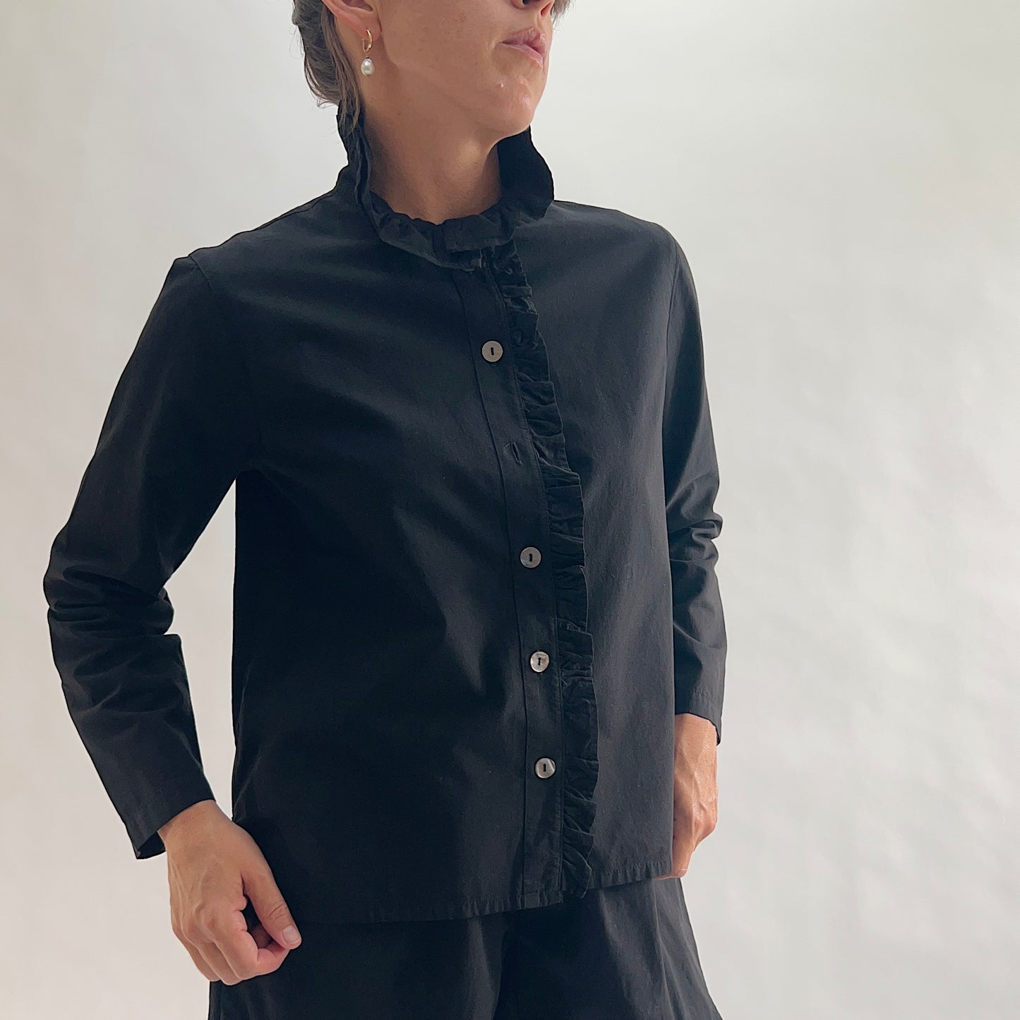Three-quarter front view of model wearing Eleven Stitch black blossom collar shirt, showing ruffled blossom collar, ruffled button placket with shell buttons, long sleeves with cuffs, relaxed boxy fit, curved hem, and black color, styled with matching black wide-leg pants