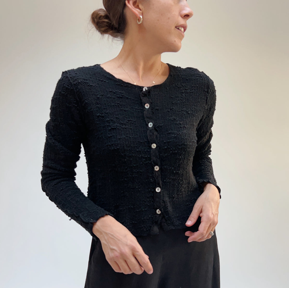 Front view on a model of a black textured knit crop cardigan with a round neckline and light buttons down the front.
