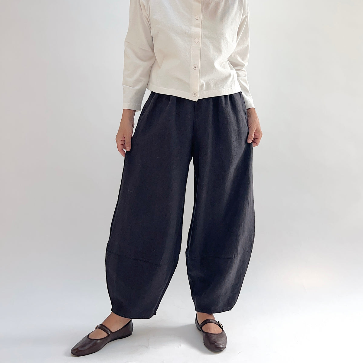 Model wearing the Bryn Walker Heavy Linen Oliver Pant in Fig, angled front view showing barrel-leg silhouette, elastic waistband, side pocket, and tapered ankle in deep fig heavy linen, styled with cream button-up and brown Mary Jane flats
