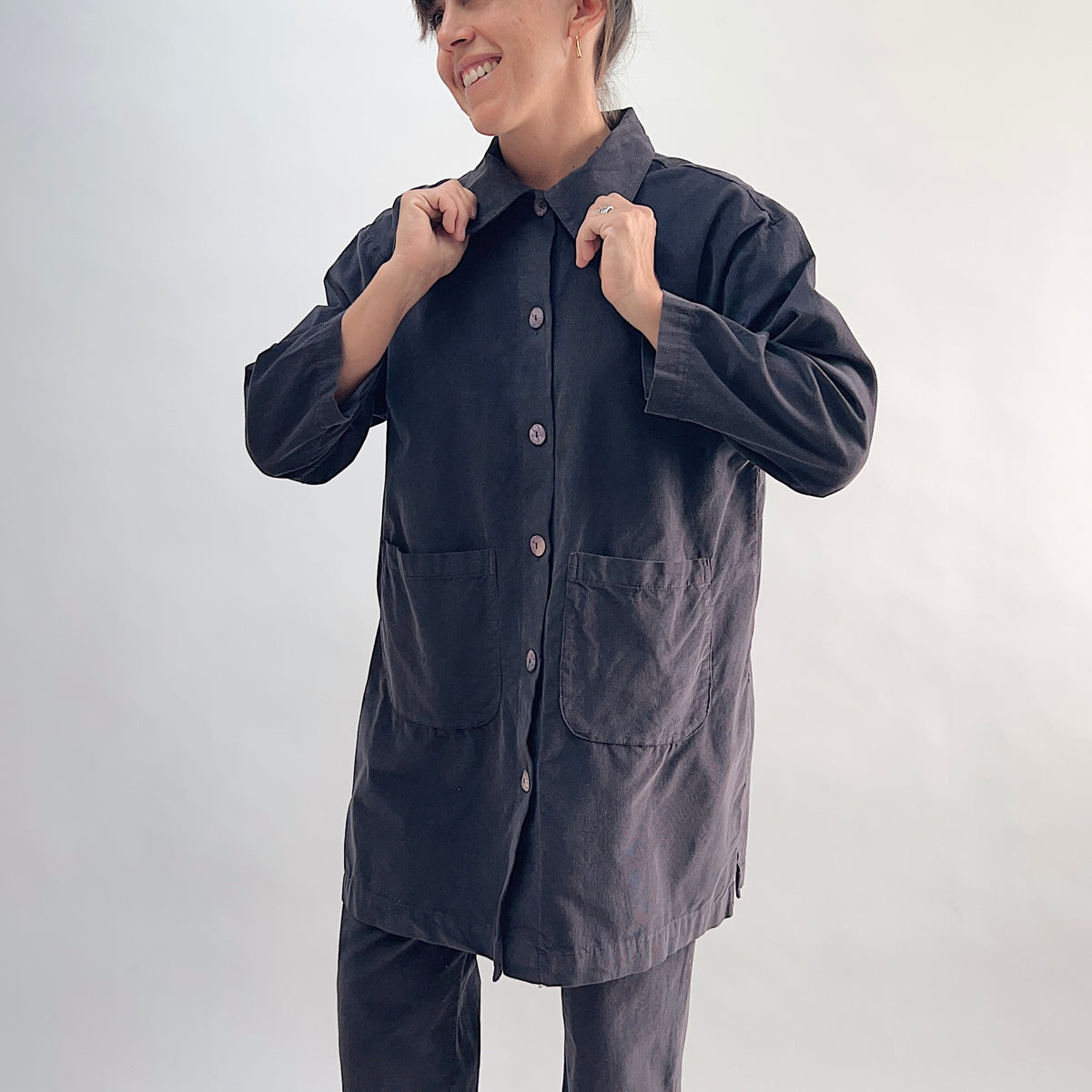 Model adjusting collar of the Bryn Walker Corduroy Car Coat in Fig, front view highlighting rounded point collar, mother of pearl button closure, two large patch pockets, 3/4 sleeves, side hem slits, and deep fig pinwheel corduroy, styled with matching corduroy pants
