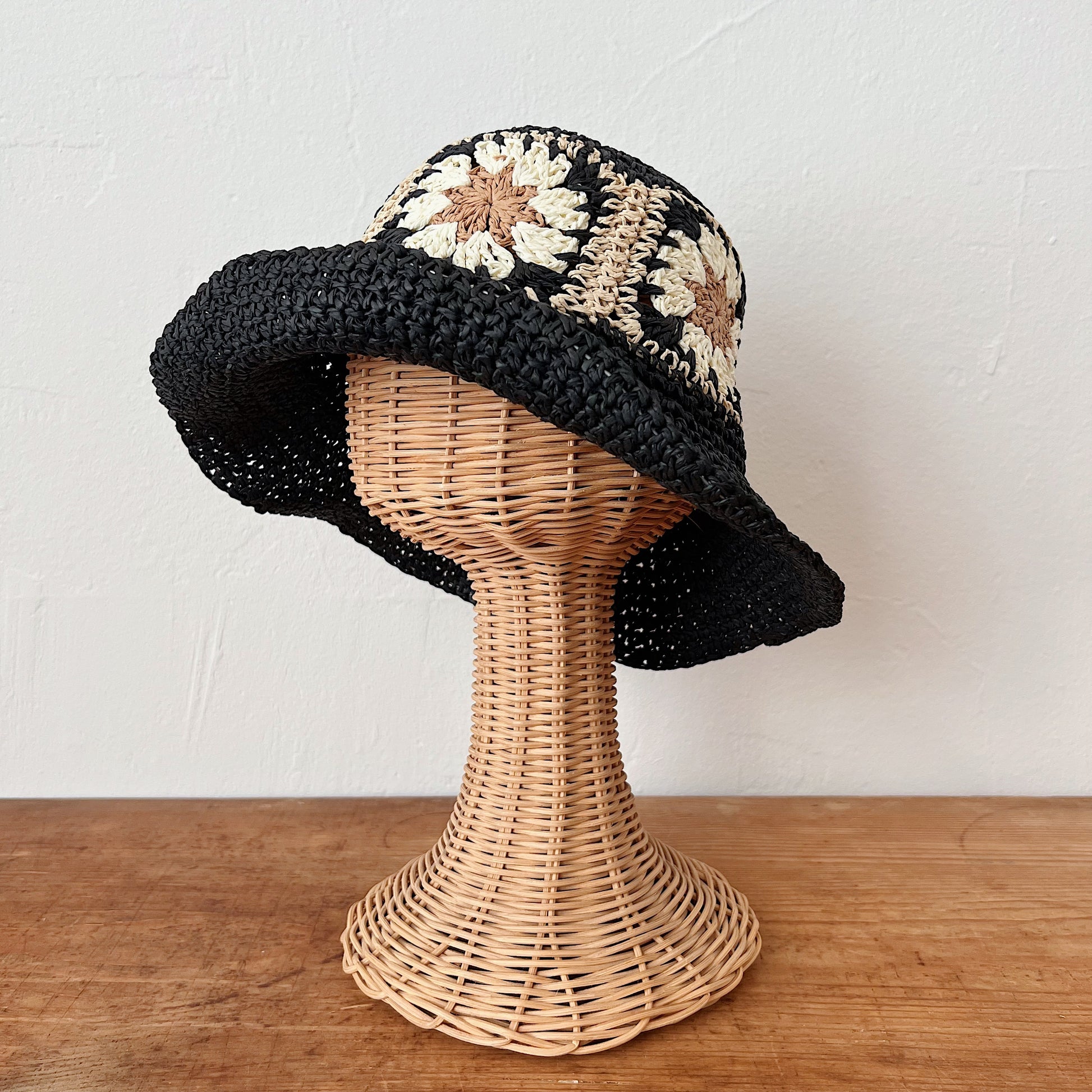 Black paper crochet bucket hat with cream and tan floral granny square band, shown on a wicker hat stand — side angle