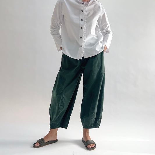 Full front view of model wearing Eleven Stitch konbu snap doreen pant with hands in pockets, showing elastic waistband, wide balloon leg tapering to snap hem at ankle, and deep forest green color, styled with white blossom collar shirt and dark slide sandals