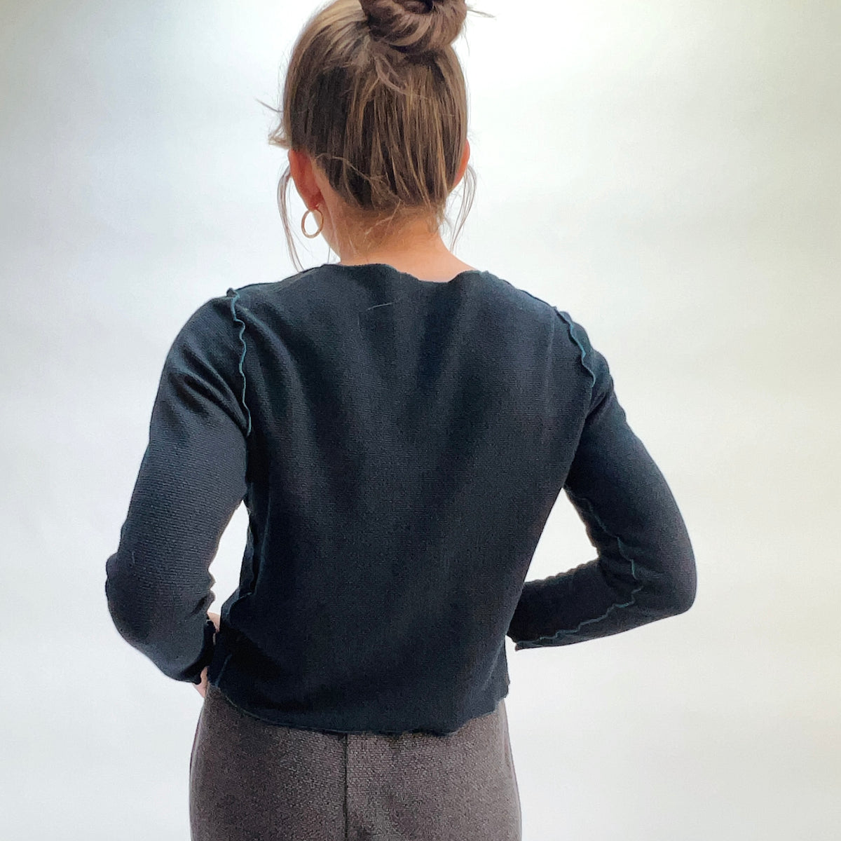Model wearing the Cut Loose Marled Knit Cropped Cardigan in Etan, back view with hair in bun and hands behind back, showing scalloped seam detail at shoulders and cropped straight hem of deep teal marled knit cardigan, styled with a dark textured skirt
