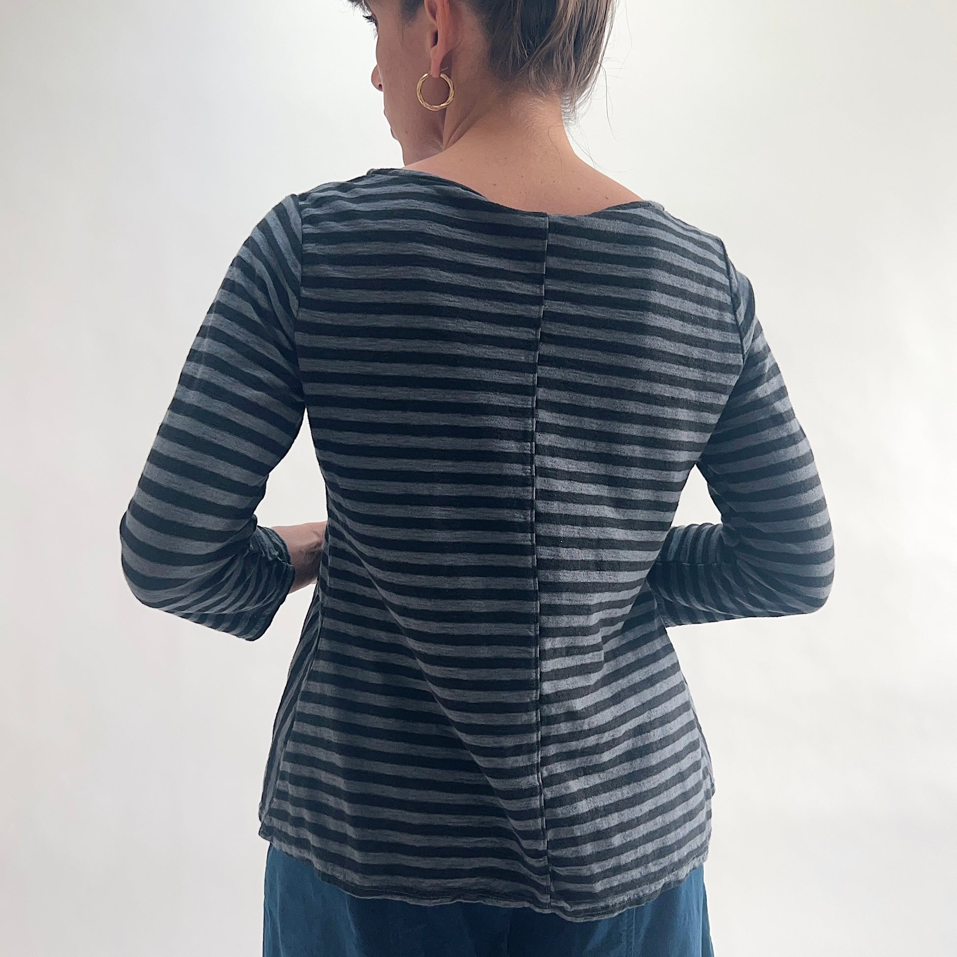 Back view of model wearing a charcoal and gray stripe A-line boatneck top with 3/4 sleeves, showing the center back seam, wide boatneck, and flared A-line silhouette, styled with dark blue wide-leg pants.
