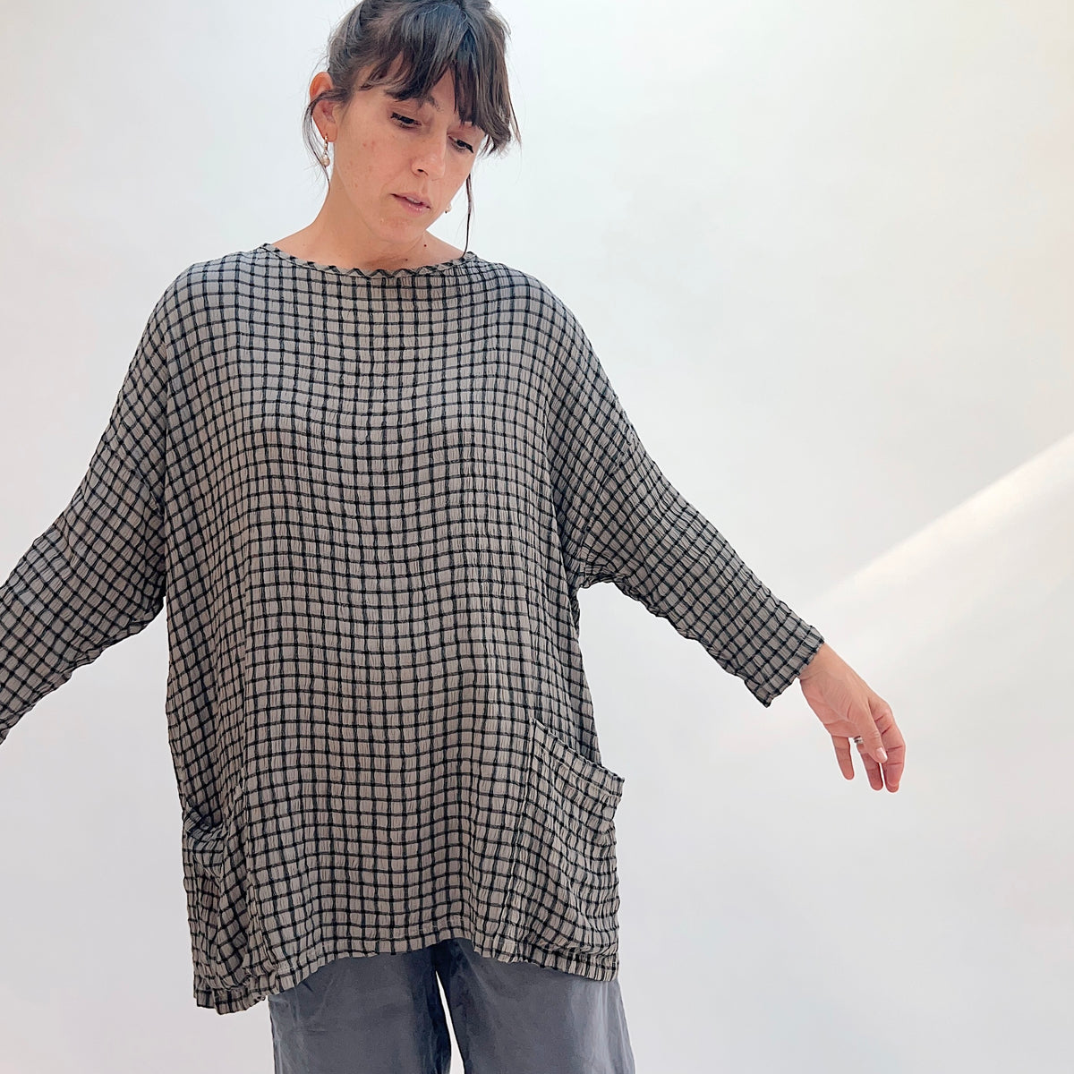 Back view of squirrel grid-check crinkle-texture one-size pocket tunic showing scoop back neckline, long sleeves, relaxed boxy silhouette, and side patch pocket, hair in bun with pearl drop earring, styled with dark grey wide-leg pants
