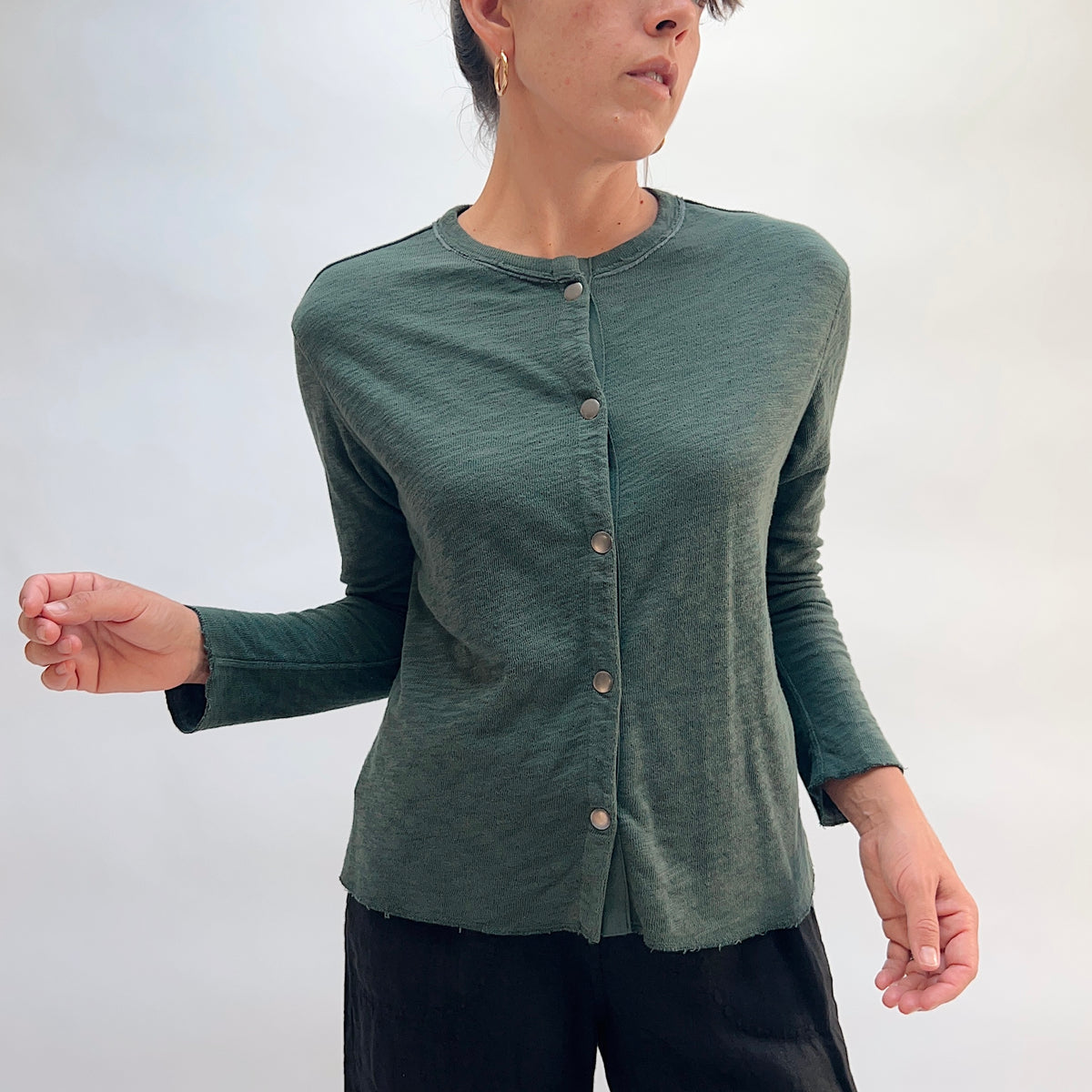 Model wearing the Cut Loose Snap Front Cardigan in Konbu, front view with arms out, showing fitted silhouette, snap button closure, and raw hem in deep teal green cotton

