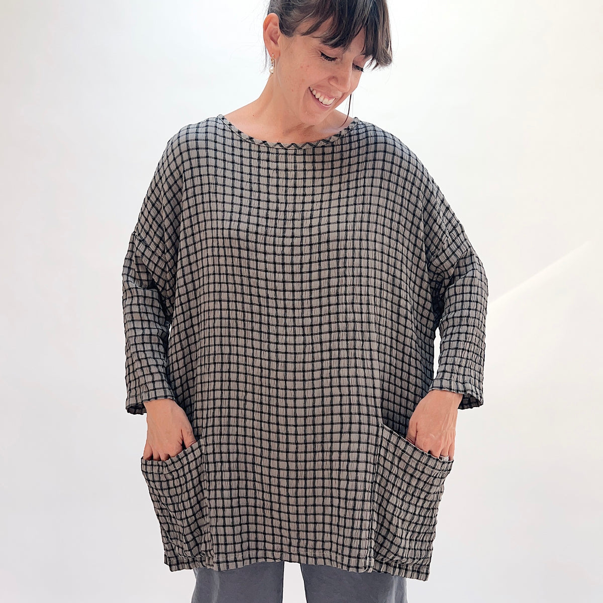 Front view of squirrel grid-check crinkle-texture one-size pocket tunic showing scoop neckline with contrast trim, long sleeves, and two front patch pockets, styled with grey wide-leg pants
