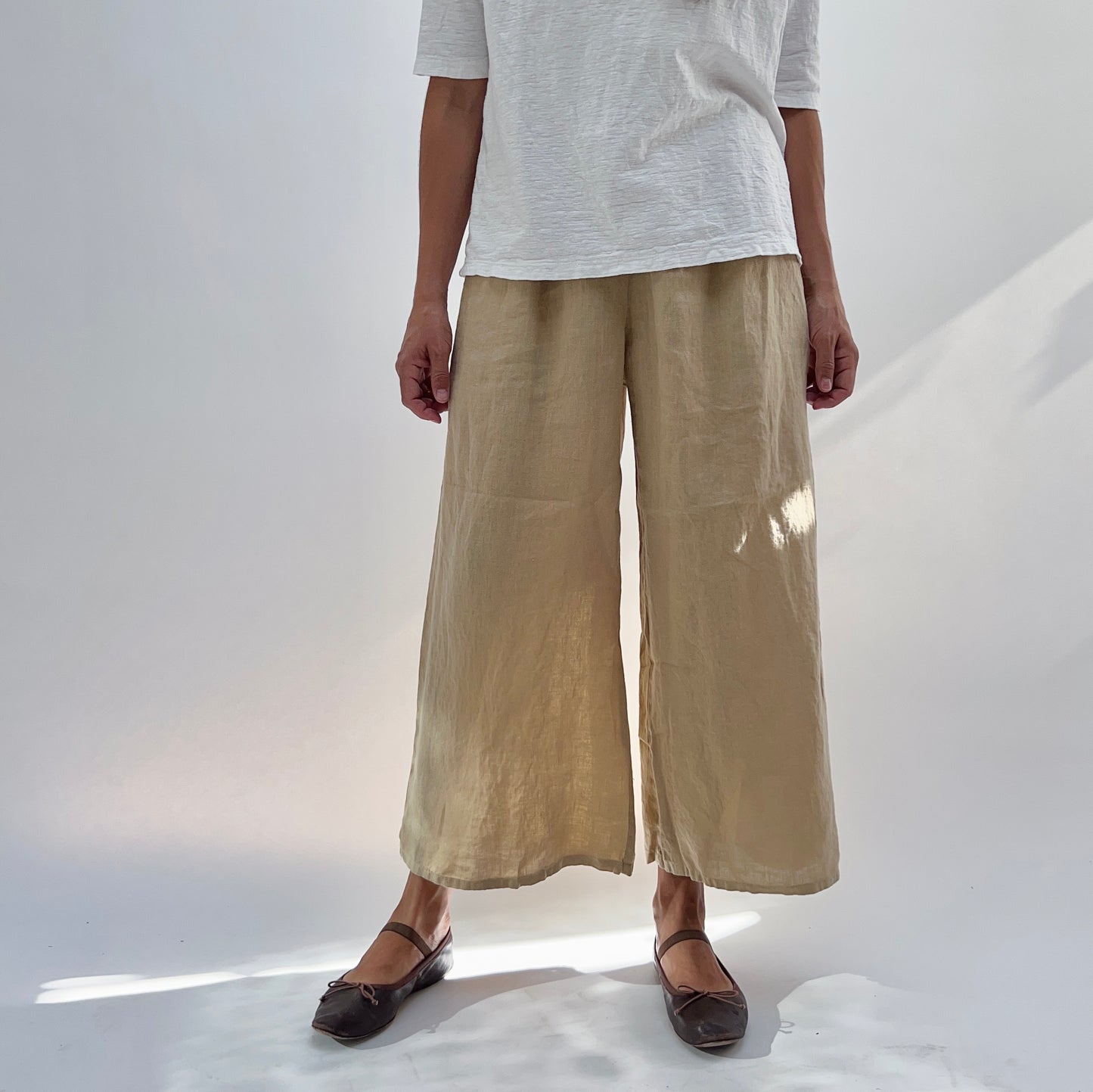 A person standing wearing beige full crop linen pants with an elastic waistband and two pockets, paired with a white top.