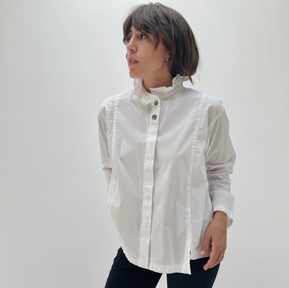 Model wearing the Eleven Stitch Tuxedo Shirt in Chalk, three-quarter front view, white cotton tuxedo shirt with ruffled collar, pintuck placket, and shell buttons, styled with black pants
