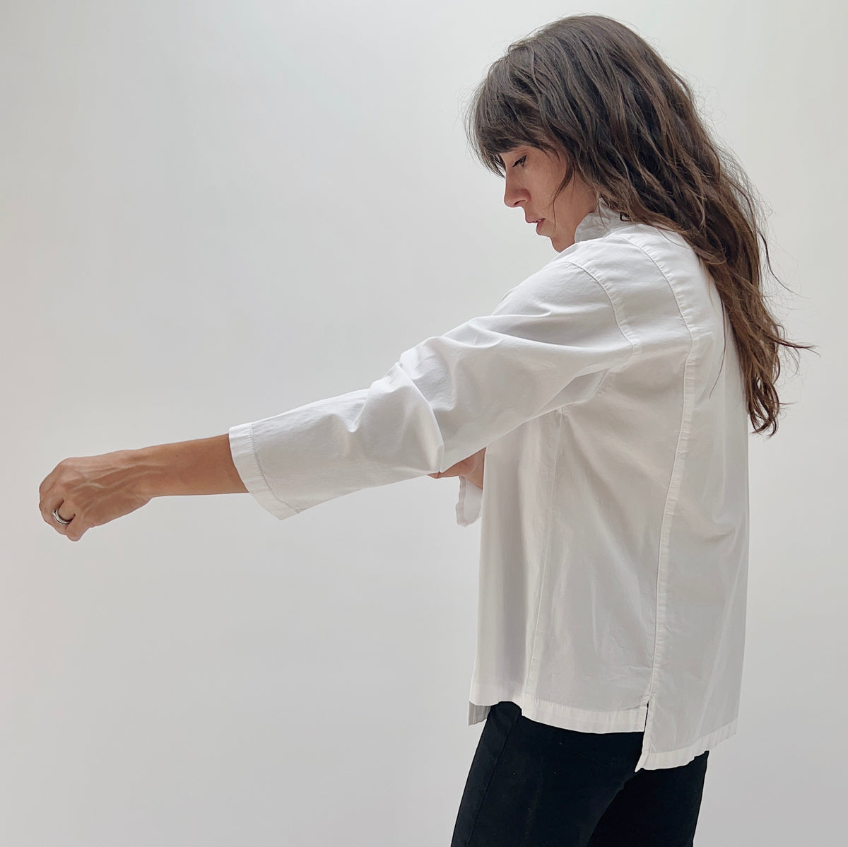 Model wearing the Eleven Stitch Tuxedo Shirt in Chalk, side profile view with arm extended, showing relaxed boxy silhouette and curved hem of white cotton tuxedo shirt
