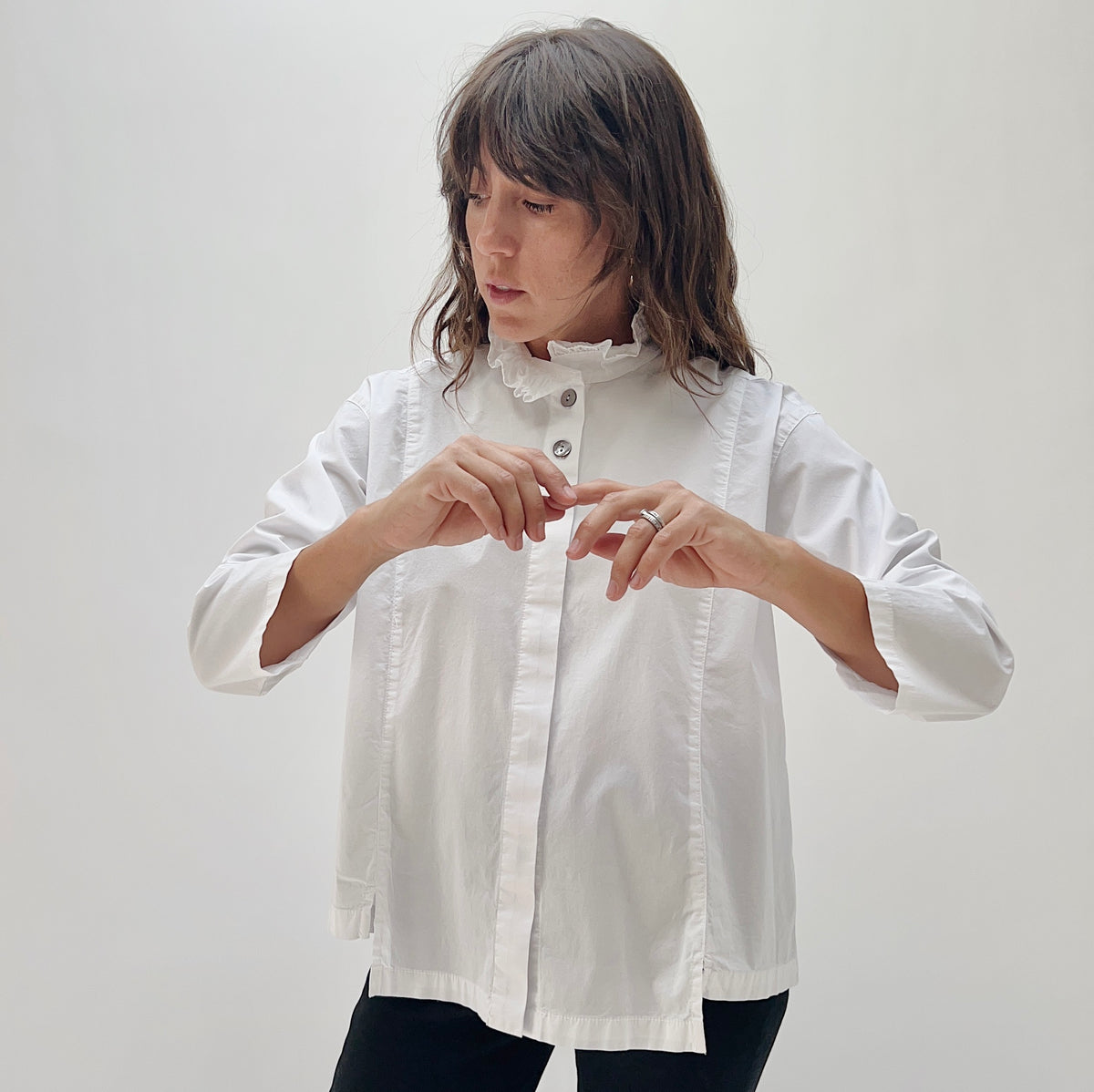 Model wearing the Eleven Stitch Tuxedo Shirt in Chalk, close-up three-quarter view, fingering shell buttons on white cotton ruffled-collar button-down shirt, styled with black pants
