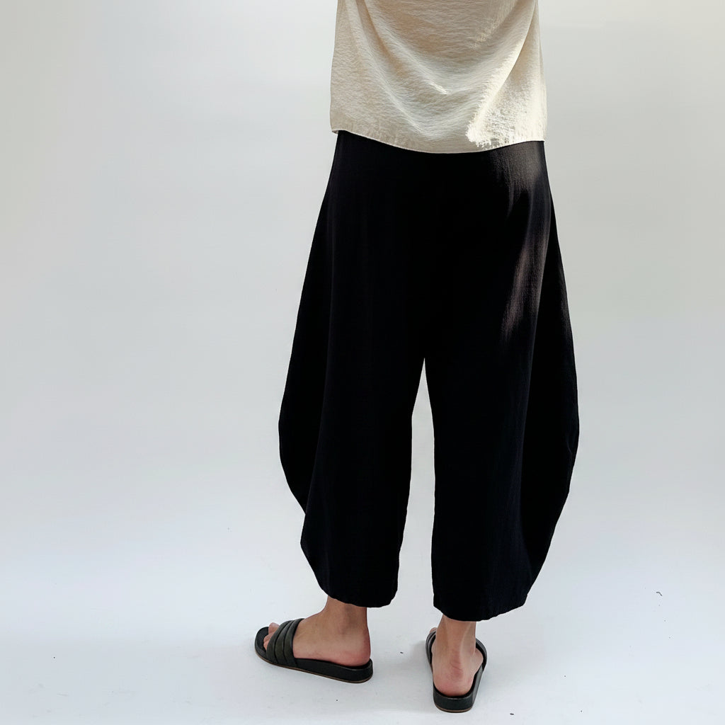 A person wearing loose, voluminous harem-style pants in a light beige and gray textured fabric, paired with a cream top and black slide sandals, shown from the waist down against a plain white studio background.