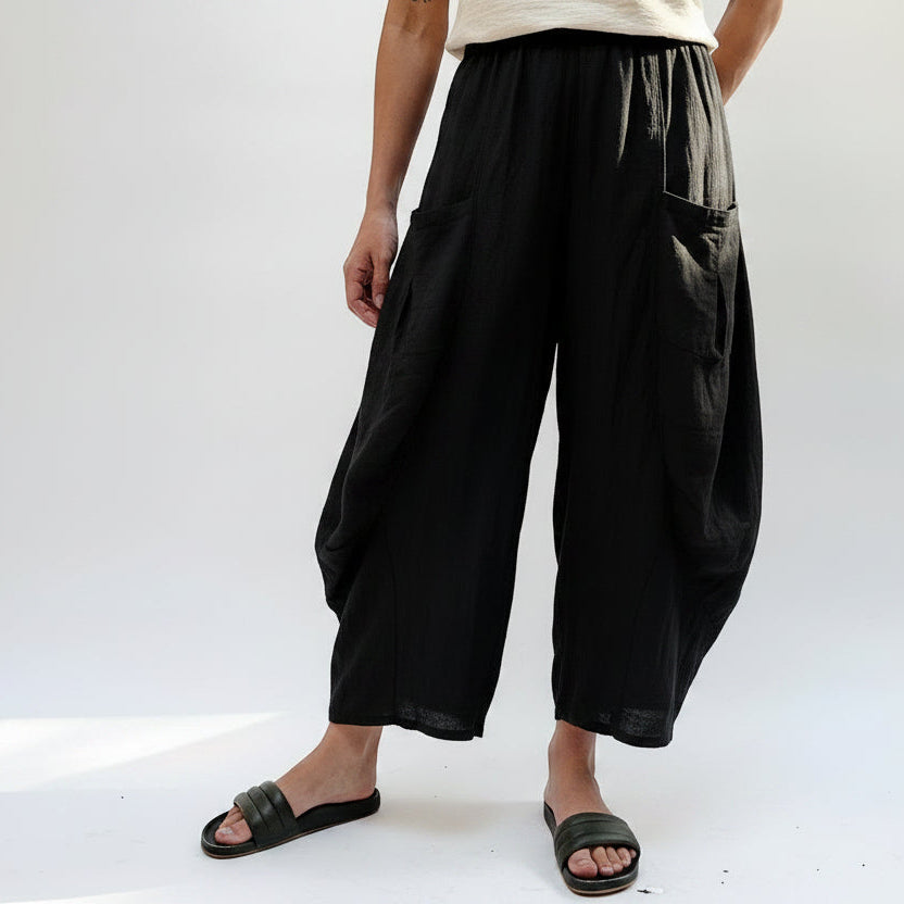 A person wearing loose, wide-leg pleated pants in a light beige and gray crinkled fabric, paired with a beige top and black slide sandals, photographed against a plain white studio background.