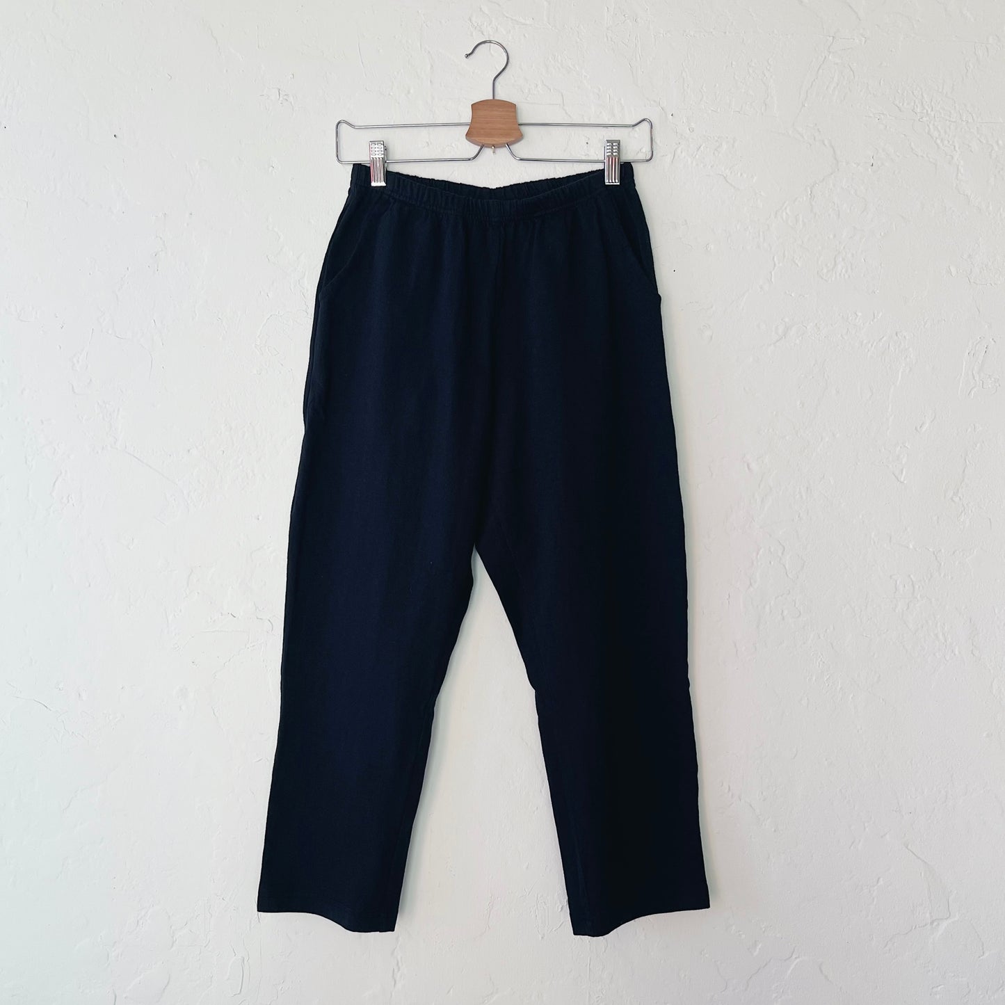 A pair of black cotton pants hanging on a hanger against a white wall.