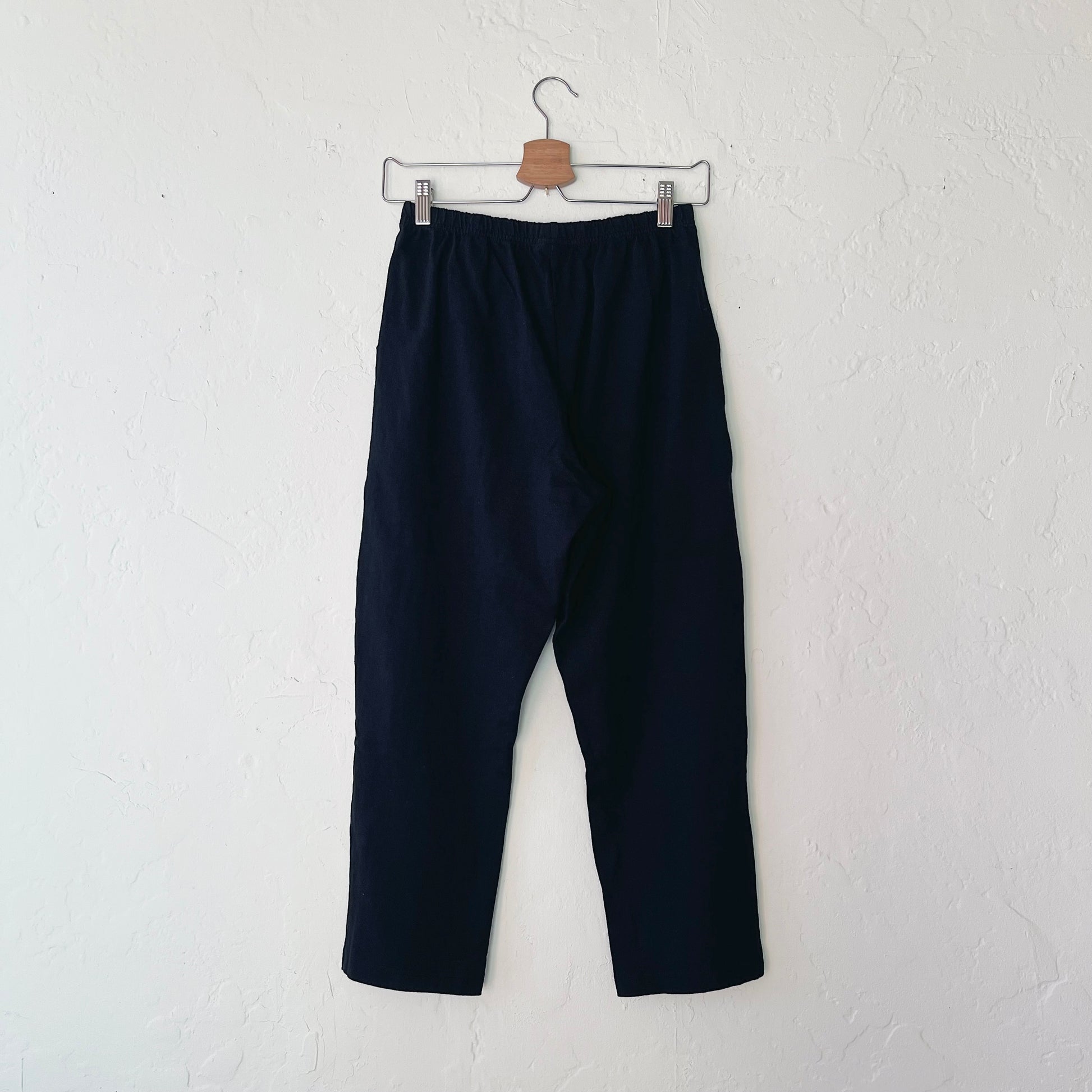A pair of black cotton pants hanging on a hanger against a white wall.