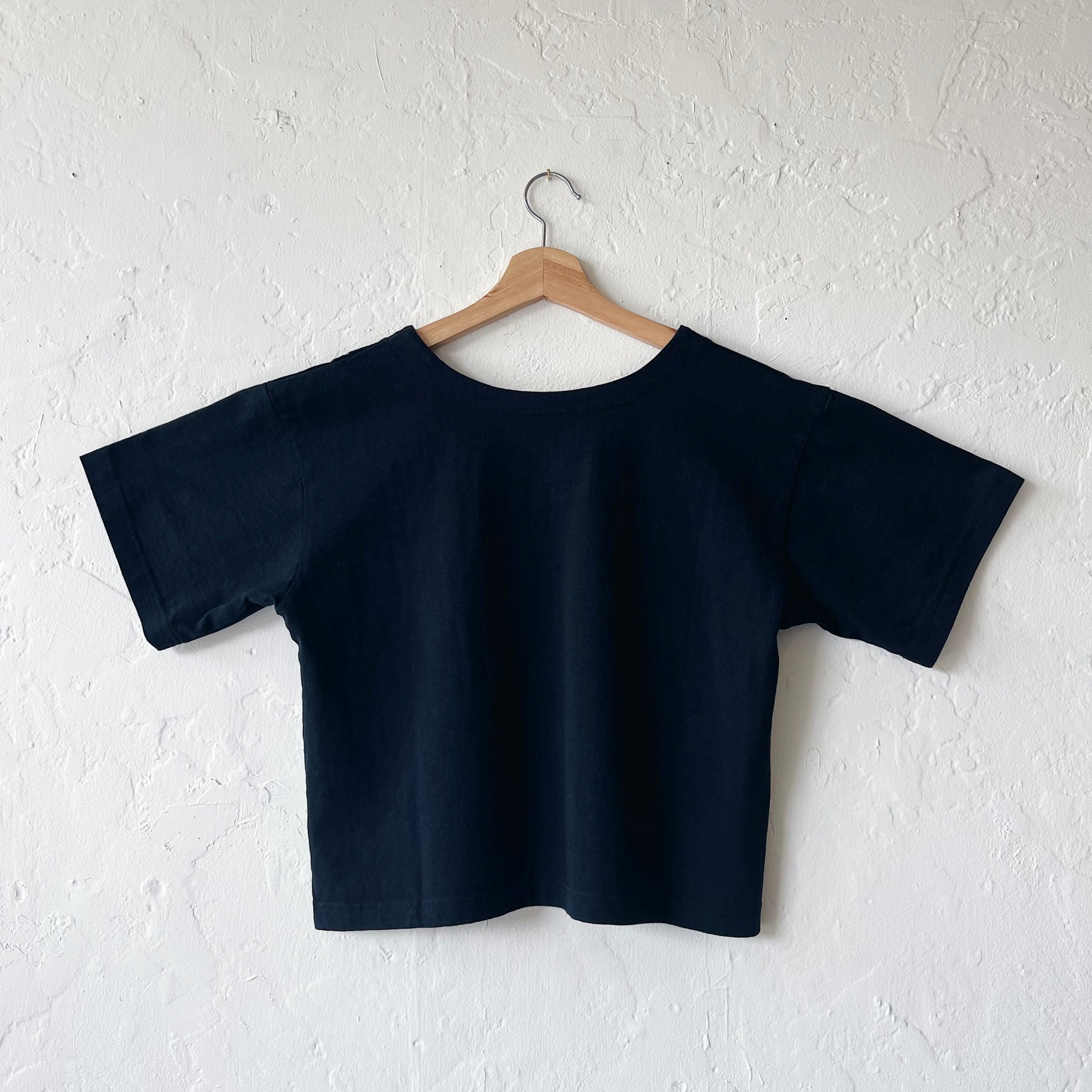 A black, cropped sleeve top with a slight v-neck hanging on a hanger against a white textured wall, viewed from the back