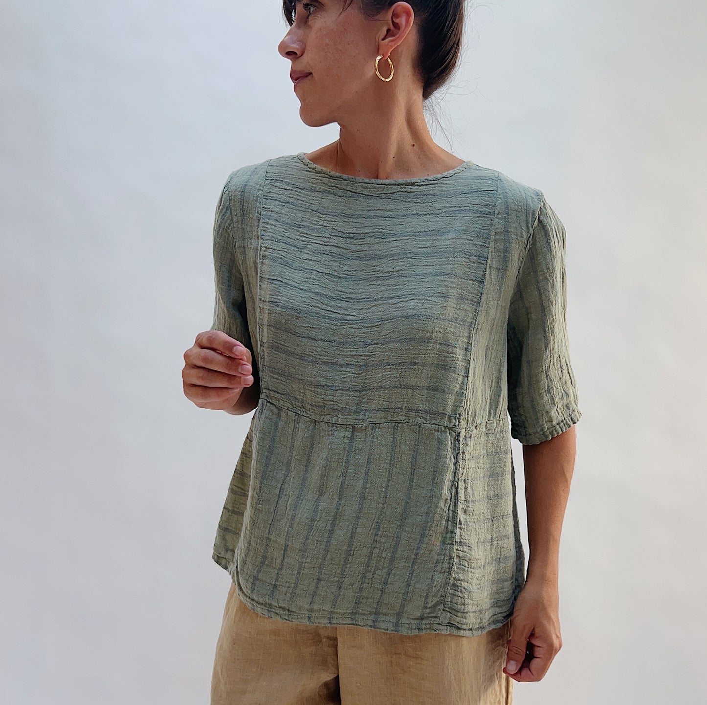 Model wearing the Seamed Boxy Striped Top in Balsam Green, a relaxed short-sleeve top with a textured stripe pattern, seamed waist detail, and crew neck, styled with tan linen pants looking off to the side