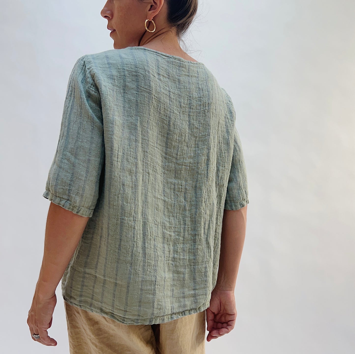 A woman shown from the back wearing a textured sage green linen short-sleeve top and beige trousers, photographed against a plain light gray studio background.