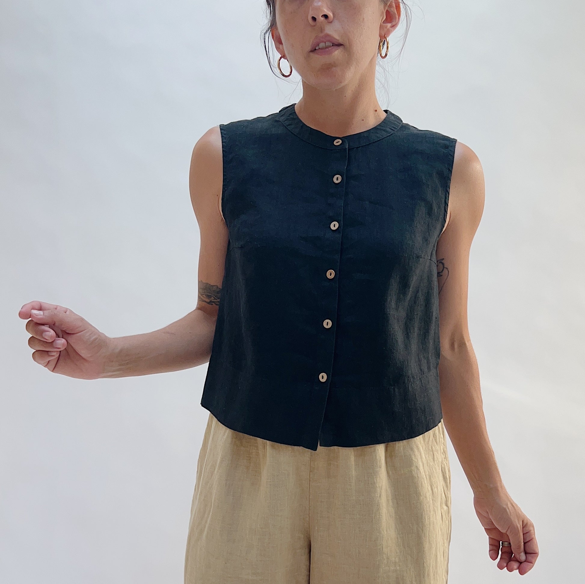 Three-quarter front view of the Button Front Tank in Black by Yuvita, showing the cropped linen silhouette, button-front detail, and collarless neckline, styled with tan linen trousers
