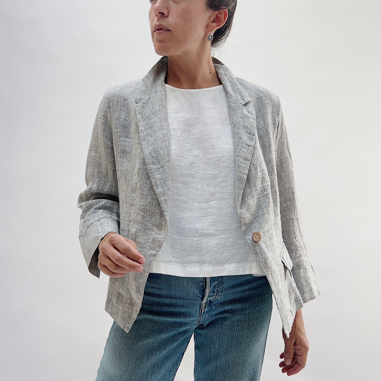 Front three-quarter view of the Linen Blazer in Grey Pinstripe by Yuvita, showing the open lapel, single-button closure, and flap pockets, styled with a white linen tee and blue jeans