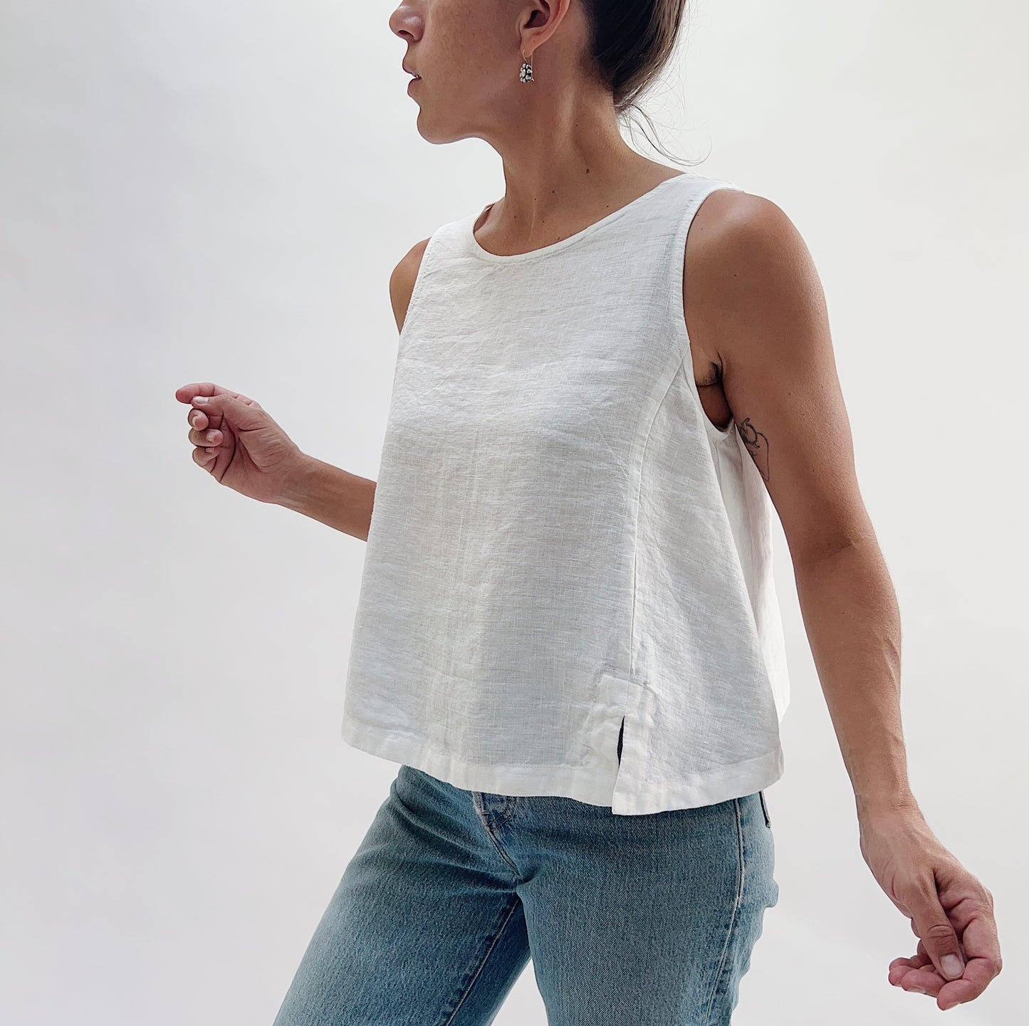 Side three-quarter view of the Button Back Tank in White by Yuvita, showing the relaxed linen drape, side slit, and cropped hem, styled with blue jeans
