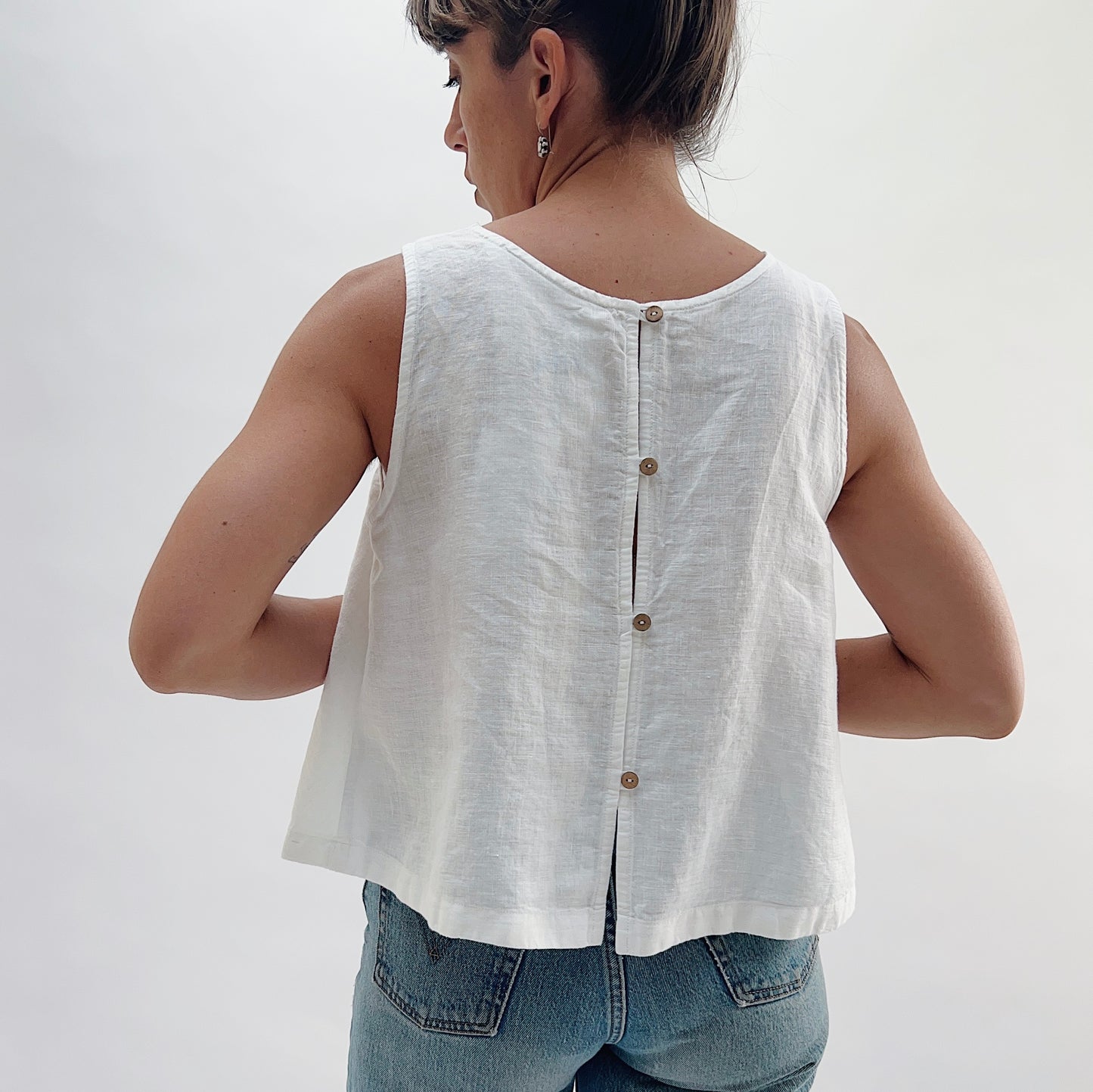 Back view of the Button Back Tank in White by Yuvita, showing the three-button back placket, scoop back neckline, and side slits in white linen, styled with blue jeans