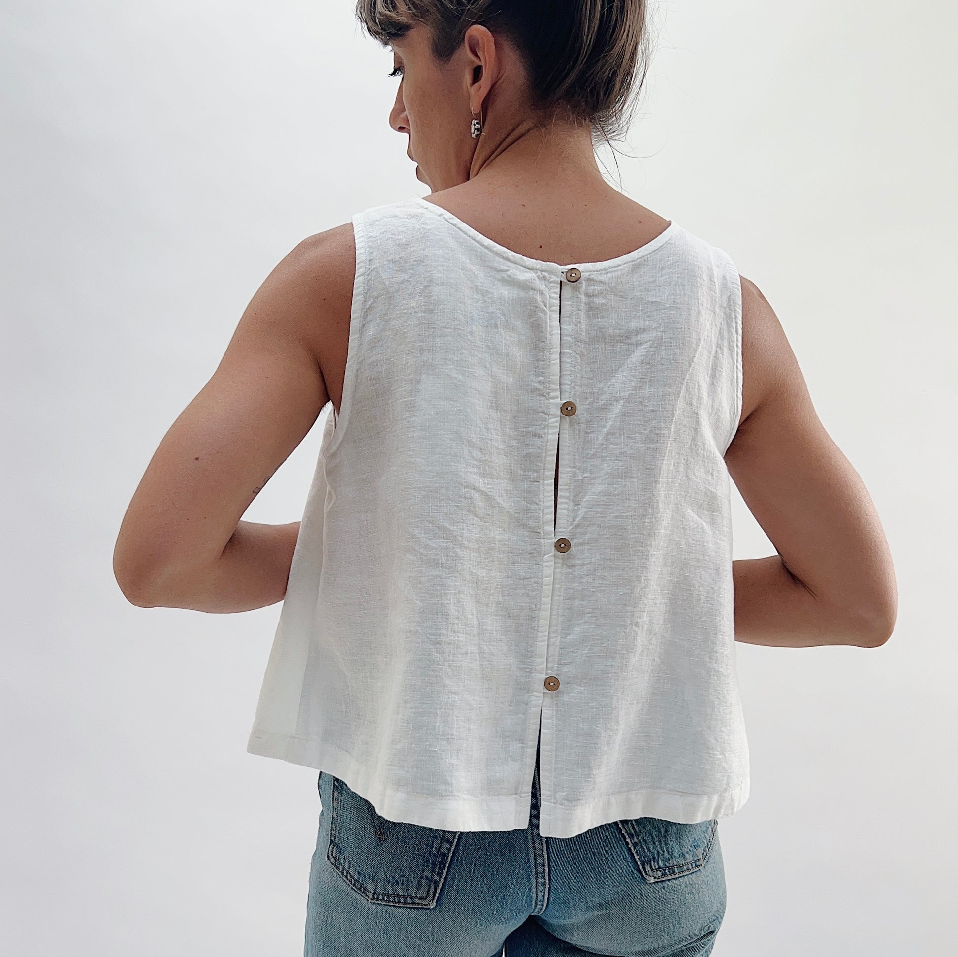 Back view of the Button Back Tank in White by Yuvita, showing the three-button back placket, scoop back neckline, and side slits in white linen, styled with blue jeans
