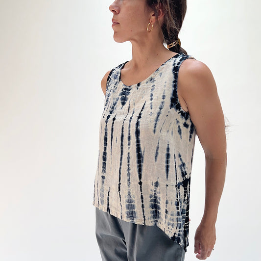 Front view of model wearing Eleven Stitch Crinkle Travel Gail Tank in indigo tie-dye, showing scoop neckline and relaxed fit worn over grey pants against a white studio background