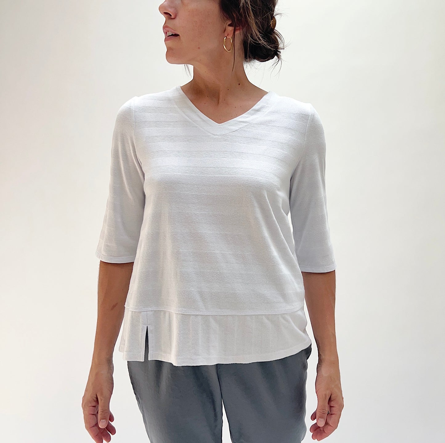 Front view of model wearing Habitat white shadow stripe elbow sleeve top, showing V-neckline, tonal stripe pattern, elbow sleeves, and layered hem with side slit
