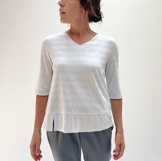 Front view of model wearing Habitat white shadow stripe elbow sleeve top, showing V-neckline, tonal stripe pattern, elbow sleeves, and layered hem with side slit