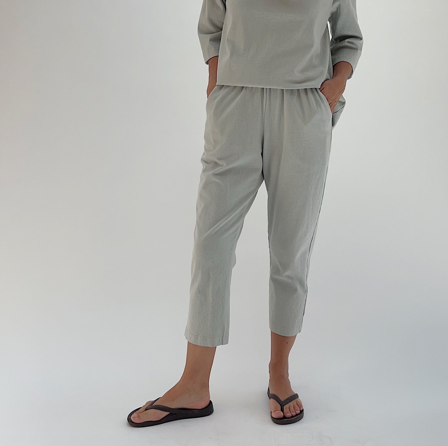 Full front view of model wearing Pacific Cotton orlaya Sunday pant, showing elastic waistband, side pockets, cropped tapered leg silhouette, and sage green color