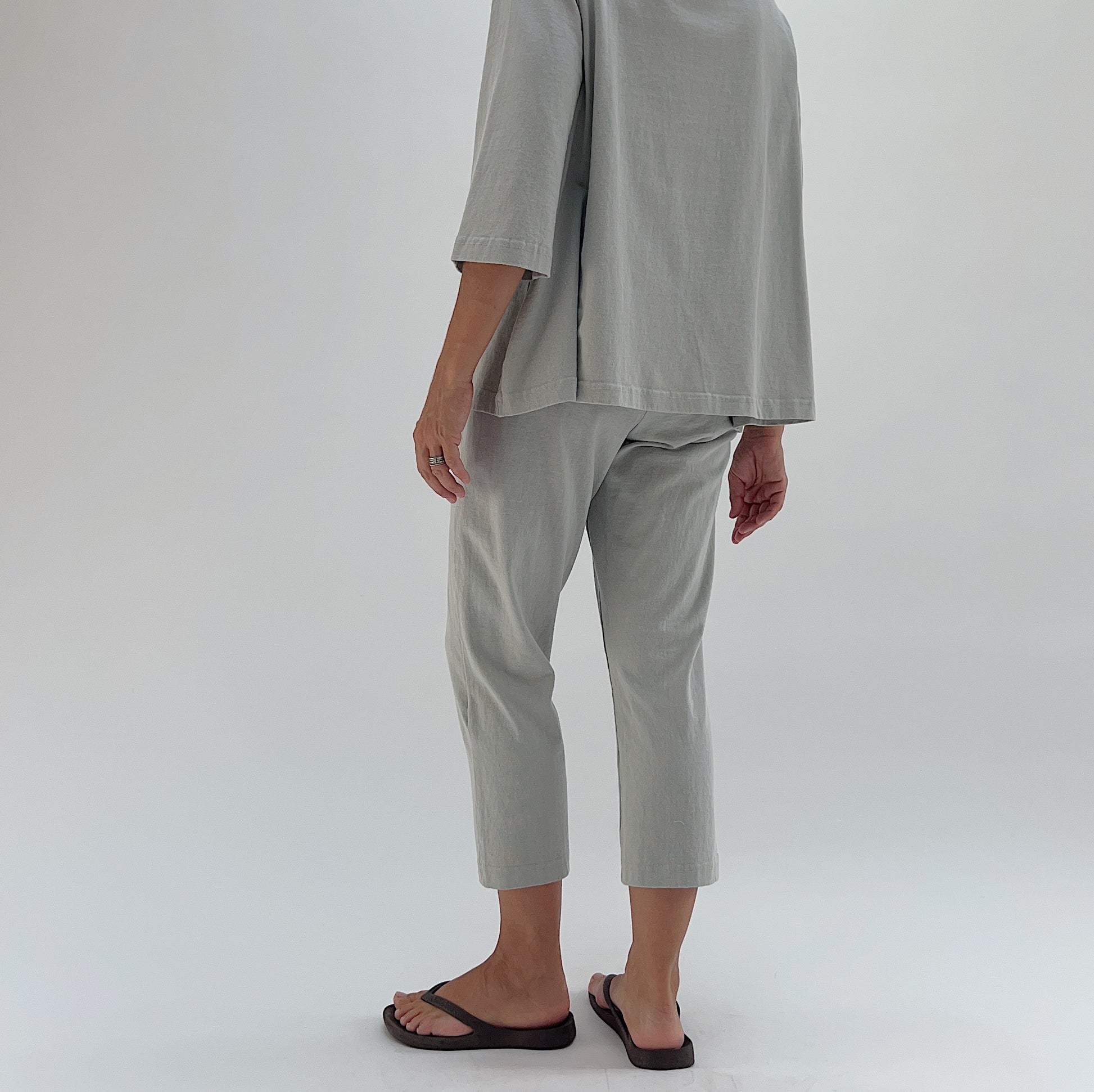 Three-quarter back view of model wearing Pacific Cotton orlaya Sunday pant, showing elastic waistband, relaxed fit through hip, cropped tapered leg, and sage green color