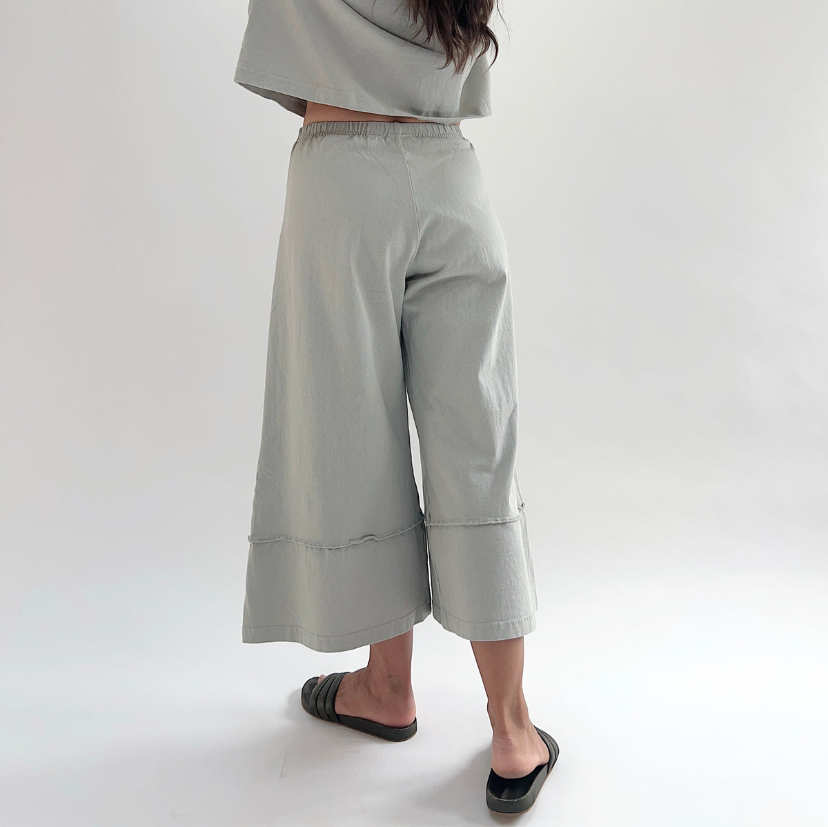Back three-quarter view of model wearing Pacific Cotton orlaya crop full pant, showing elastic waistband, wide flared leg, layered hem panel, and sage green color