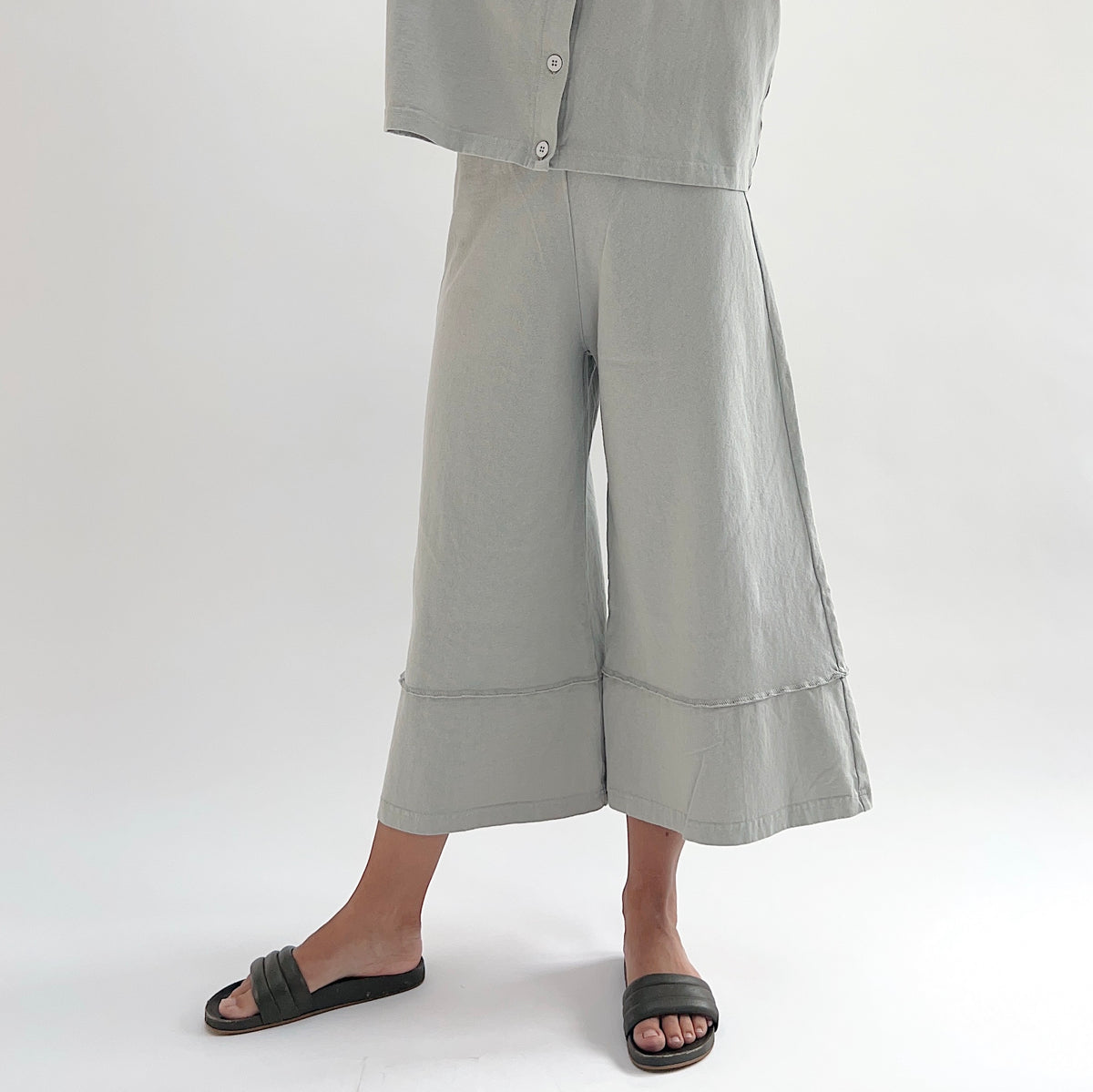 Three-quarter front view of model wearing Pacific Cotton orlaya crop full pant, showing elastic waistband, wide flared leg silhouette, layered hem panel, and cropped length