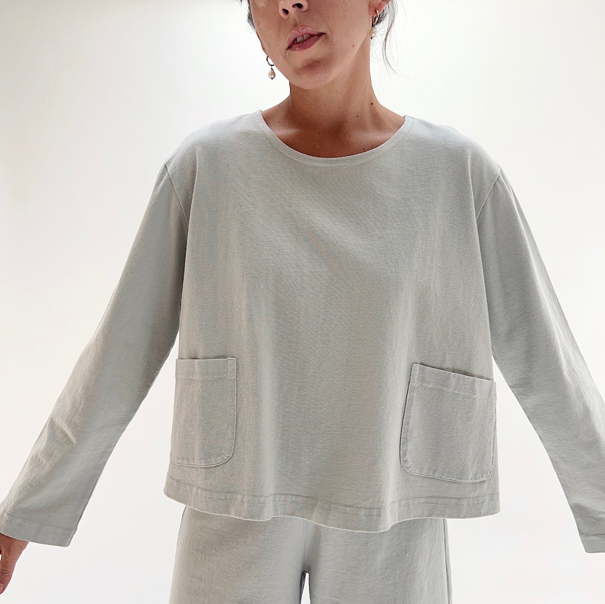 Front view of model wearing Pacific Cotton orlaya boxy shirt, showing crew neckline, two front patch pockets, long sleeves, boxy cropped fit, and sage green color
