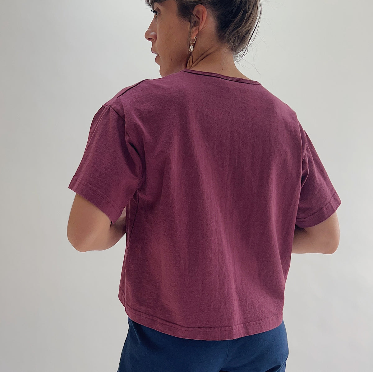 Back view of model wearing Pacific Cotton rhone crop crew tee, showing crew neckline, short cuffed sleeves, boxy cropped silhouette, banded hem, and berry plum color