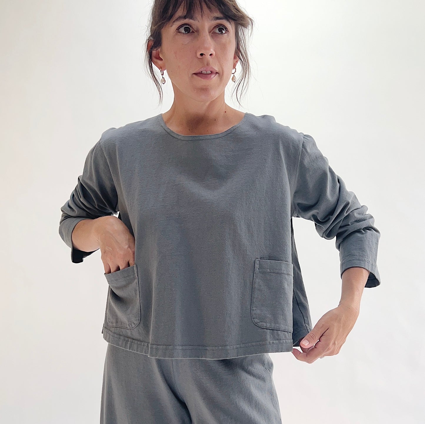 Full front view of model wearing Pacific Cotton dolomite boxy shirt with hands in pockets, showing crew neckline, two front patch pockets, long sleeves, boxy cropped fit, and washed slate grey color, styled with matching dolomite pants