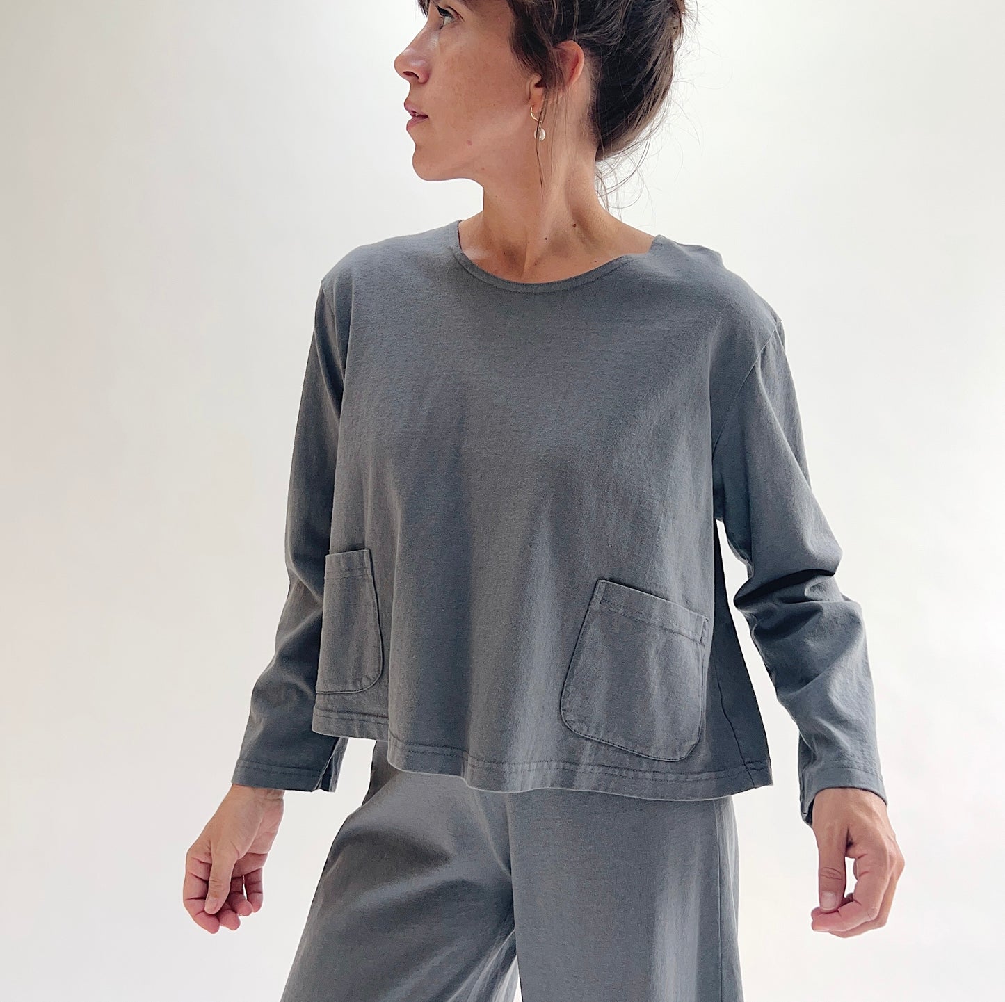 Three-quarter front view of model wearing Pacific Cotton dolomite boxy shirt, showing crew neckline, two front patch pockets, long sleeves, boxy cropped silhouette, banded hem, and washed slate grey color, styled with matching dolomite pants