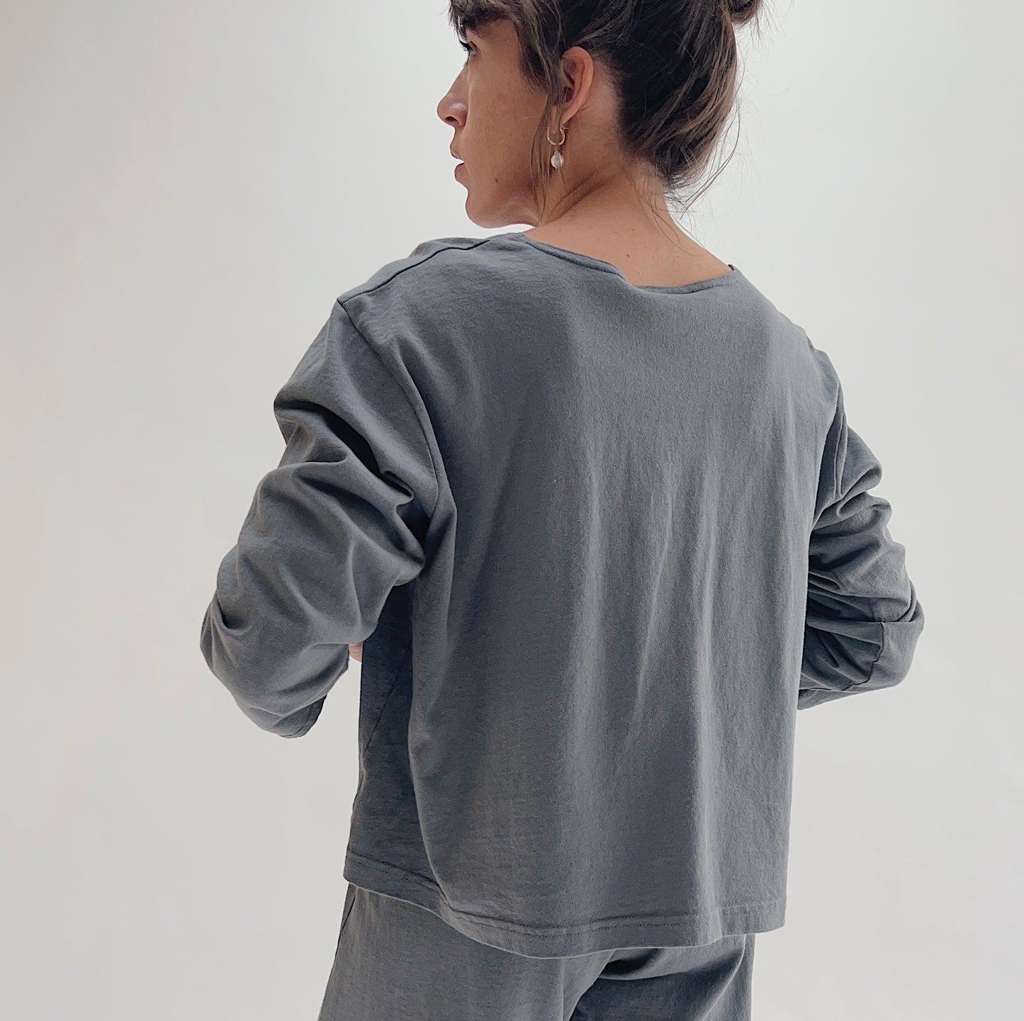 Back view of model wearing Pacific Cotton dolomite boxy shirt, showing wide back neckline, long sleeves, boxy cropped silhouette, banded hem, and washed slate grey color, styled with matching dolomite pants