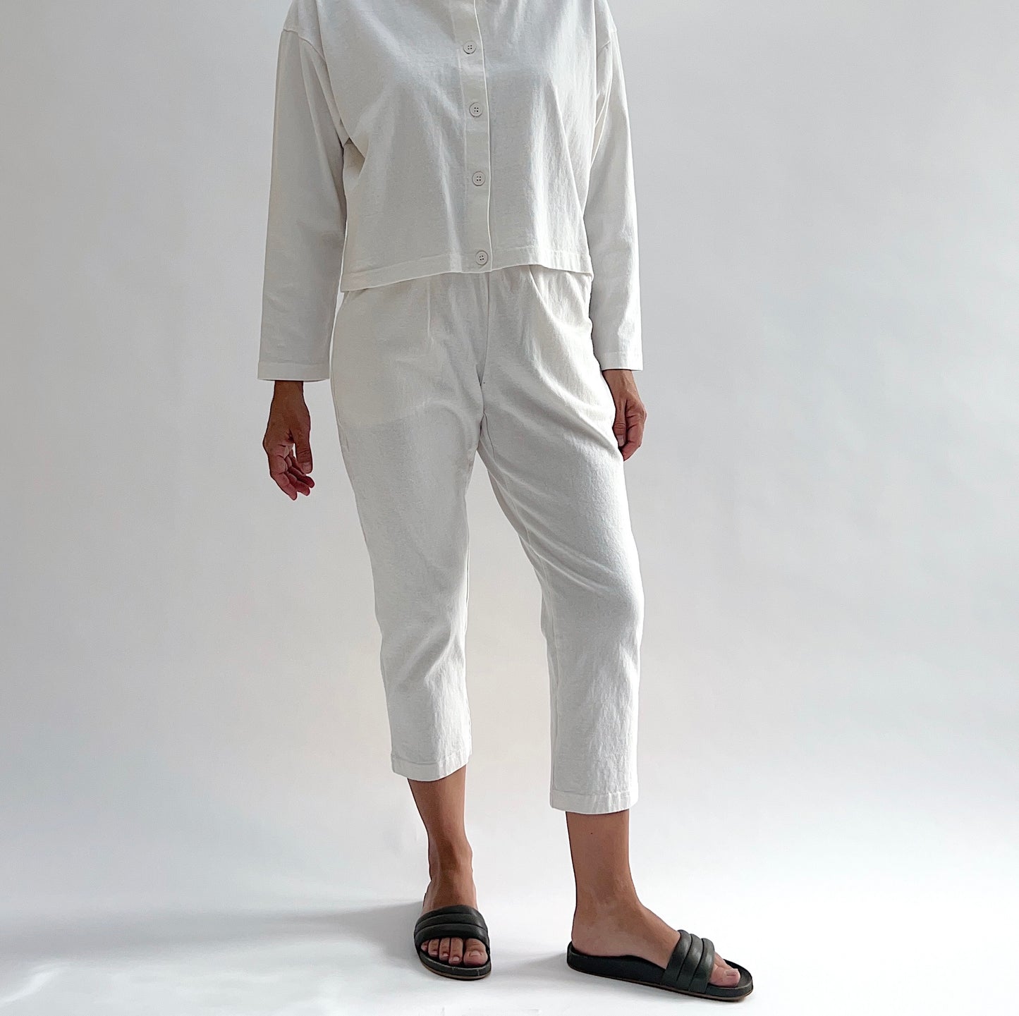 Full front view of model wearing Pacific Cotton cream sunday pant, showing elastic waistband, side pockets, cropped tapered leg, and off-white color, styled with matching cream long sleeve button front shirt and black slide sandals