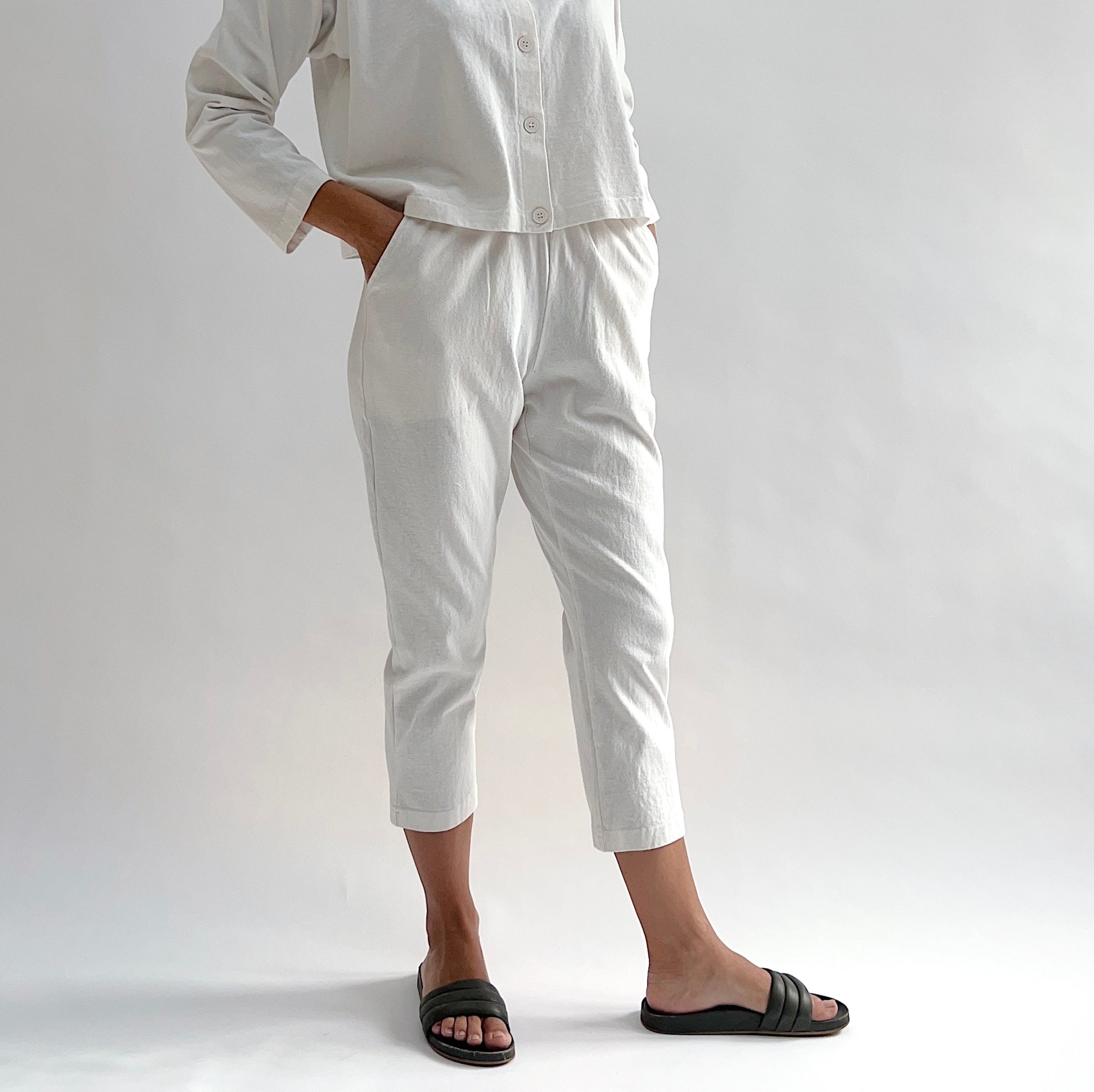 Close front view of model wearing Pacific Cotton cream sunday pant with hand in pocket, showing elastic waistband, side pocket, cropped tapered leg, and off-white color, styled with matching cream long sleeve button front shirt