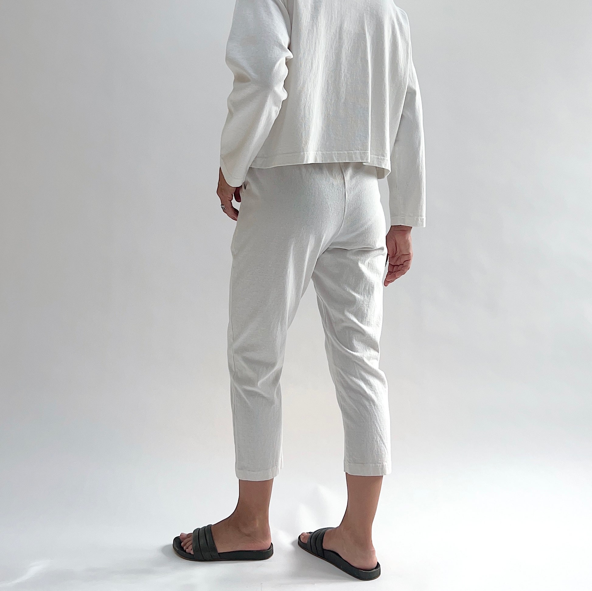 Back view of model wearing Pacific Cotton cream sunday pant, showing elastic waistband, cropped tapered leg silhouette, and off-white color, styled with matching cream long sleeve button front shirt and black slide sandals