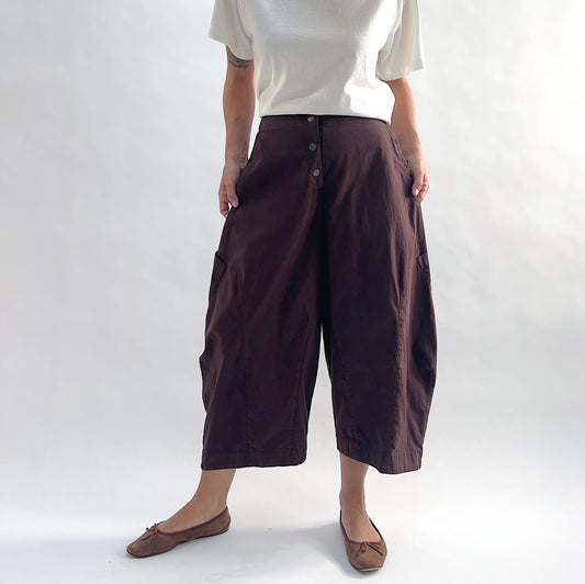 Full front view of model wearing Eleven Stitch chicory snap front pant, showing snap front waistband, wide balloon leg, large side patch pockets, gathered elastic ankle hem, and deep espresso brown color, styled with cream short sleeve tee and taupe ballet flats