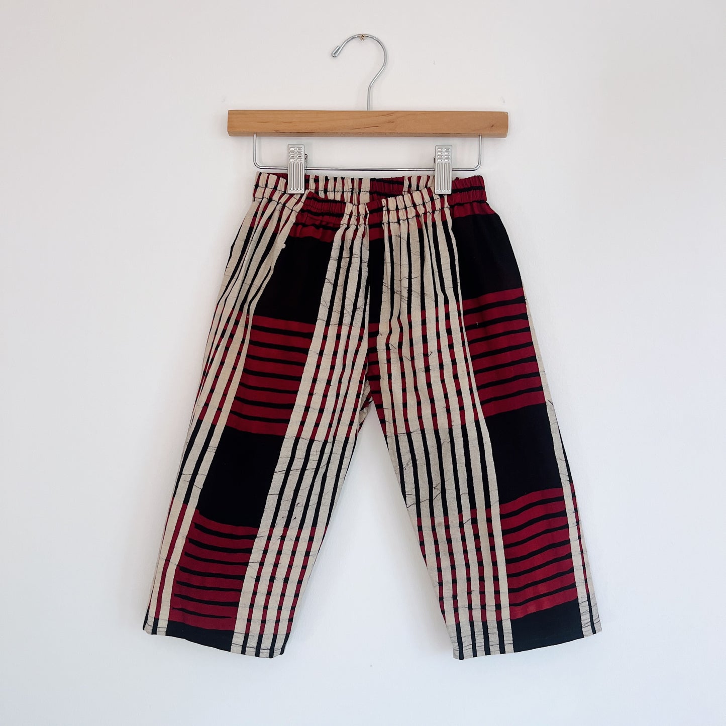 Kids lazy pants in red plaid Batiki cotton, hanging flat on a wooden clip hanger against a white wall, showing elasticated waistband, bold red, black, and cream plaid pattern, and relaxed fit