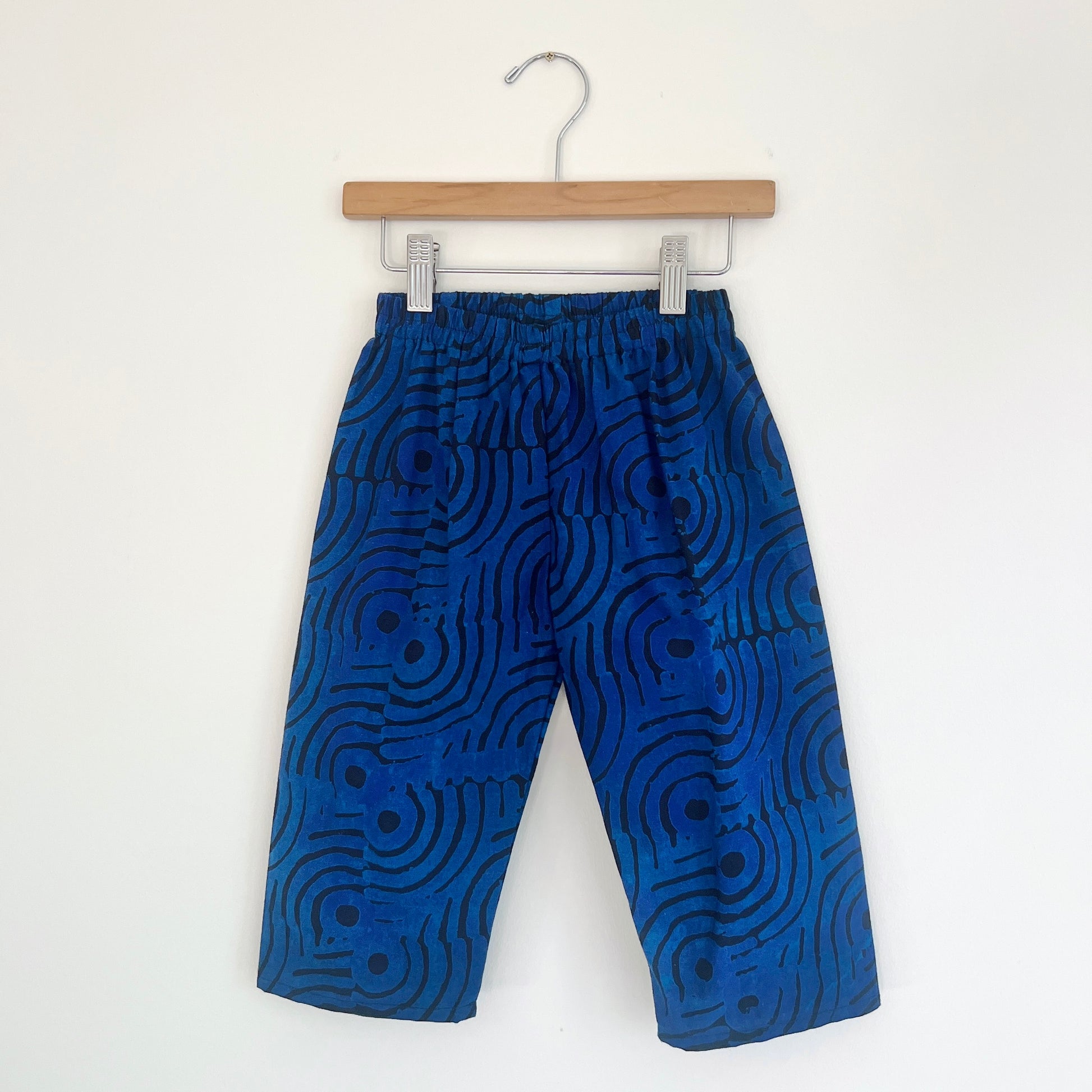 Kids lazy pants in blue waves Batiki cotton, hanging flat on a wooden clip hanger against a white wall, showing elasticated waistband, bold blue and black swirling wave print, and relaxed fit