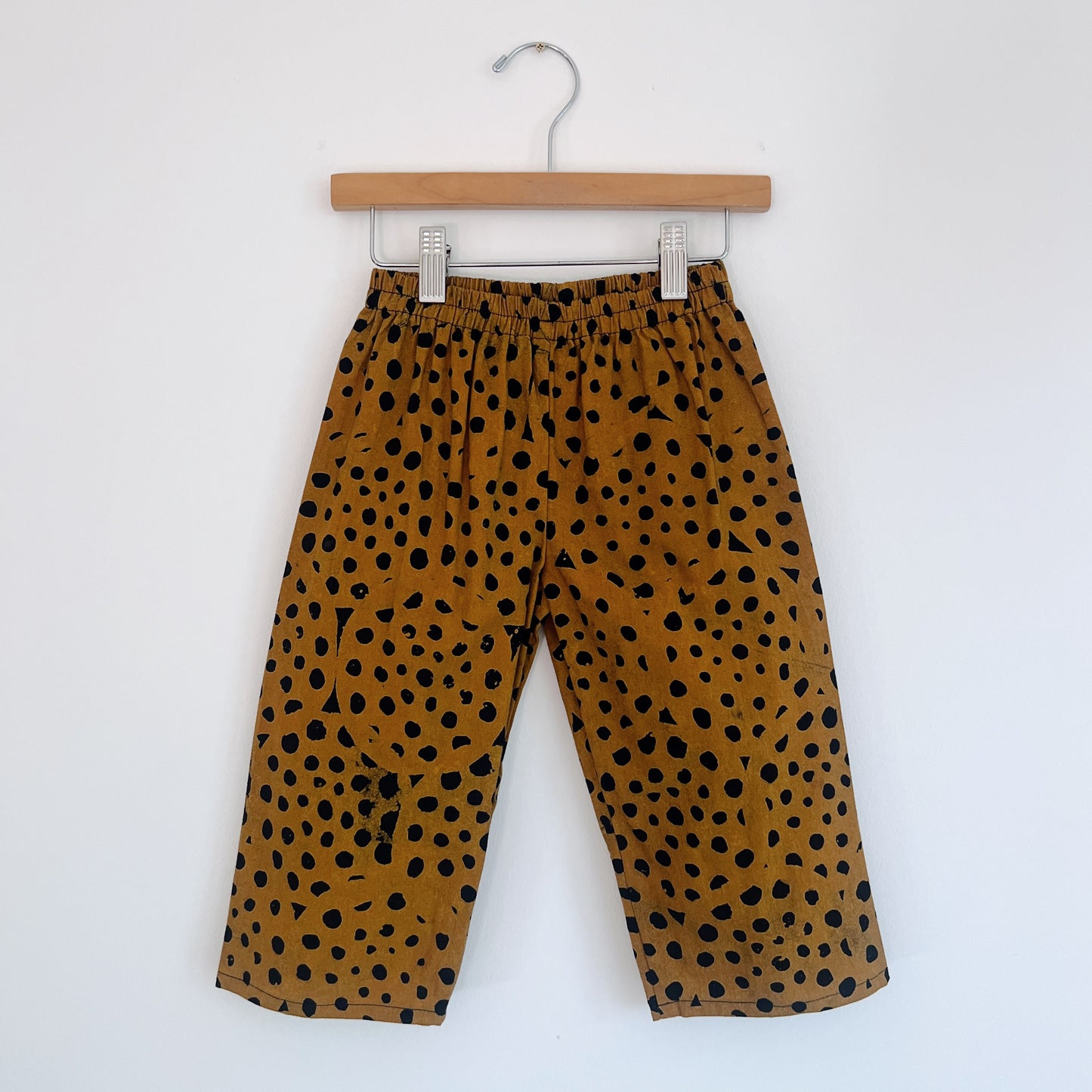 Kids lazy pants in cheetah spot Batiki cotton, hanging flat on a wooden clip hanger against a white wall, showing elasticated waistband, golden amber base with hand-printed black cheetah spots, and relaxed fit