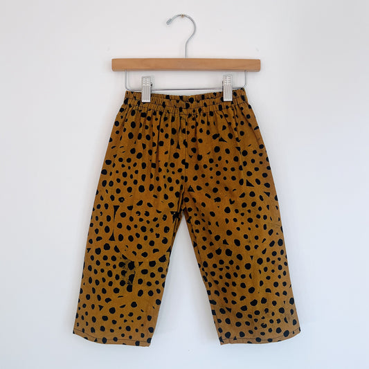 Kids lazy pants in cheetah spot Batiki cotton, hanging flat on a wooden clip hanger against a white wall, showing elasticated waistband, golden amber base with hand-printed black cheetah spots, and relaxed fit